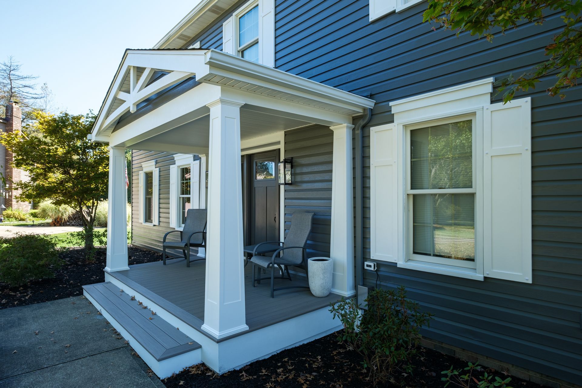 Front porch of a house with gray siding and white trim; two chairs on the porch.