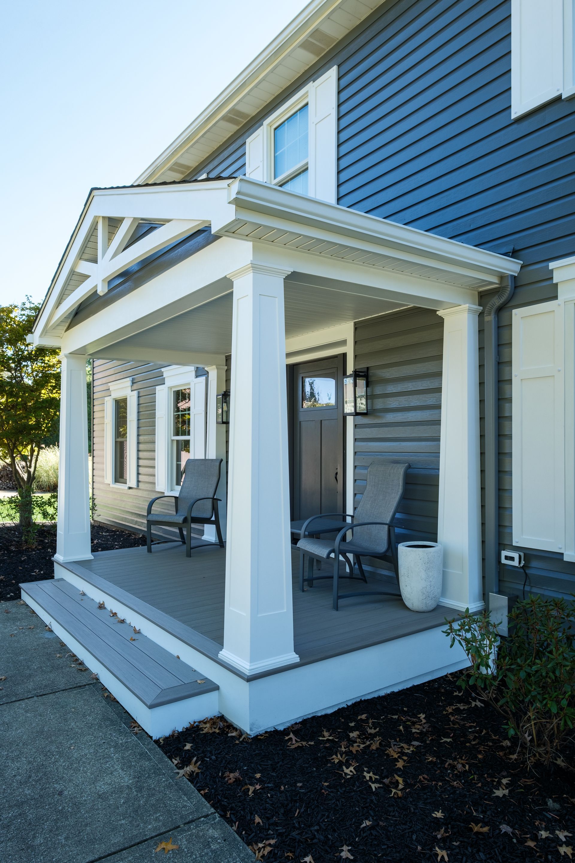 Front porch with gray siding, white columns, two chairs, and a dark door.