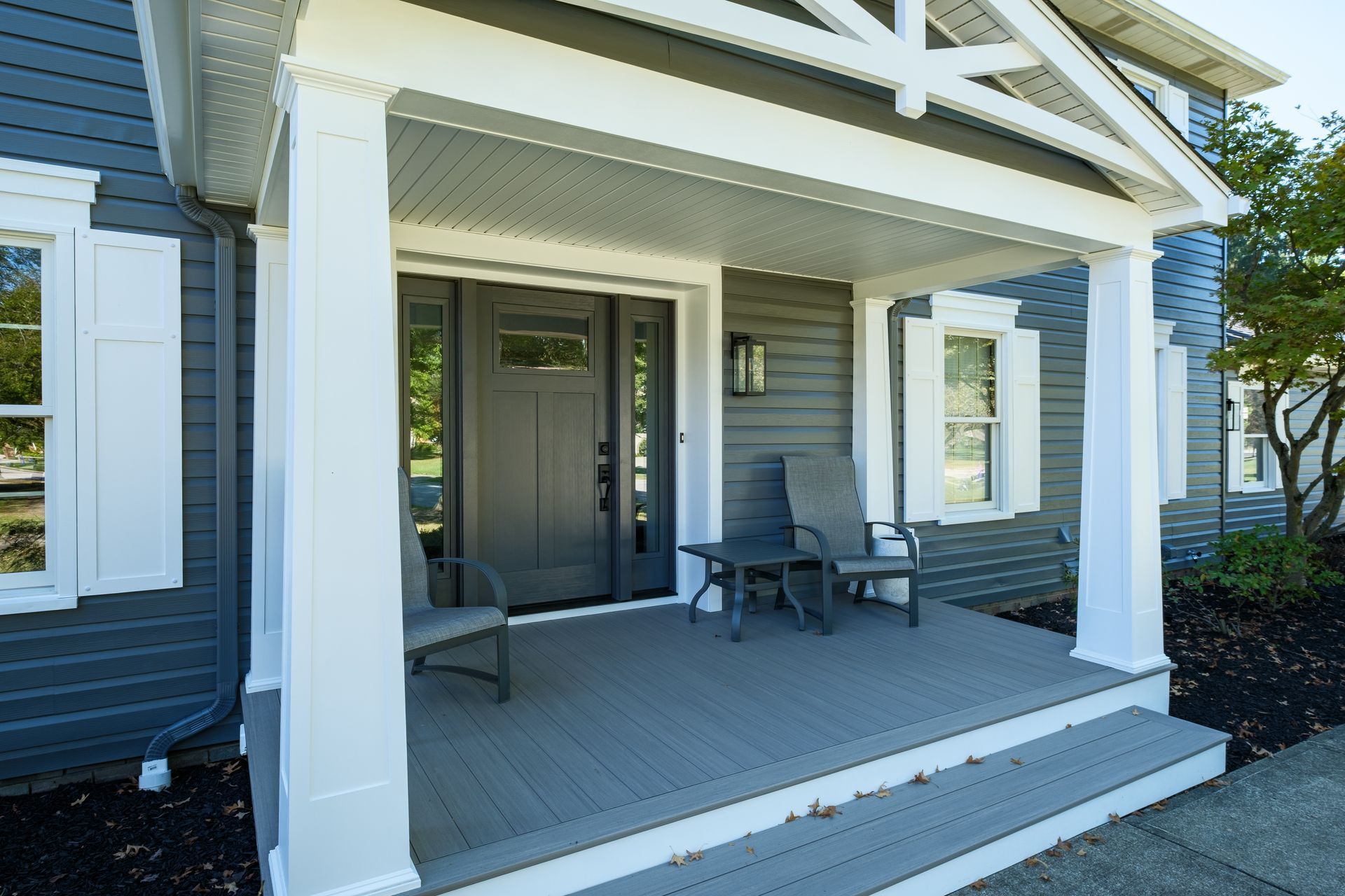 Gray house front porch with chairs and table, white columns, and a dark front door.