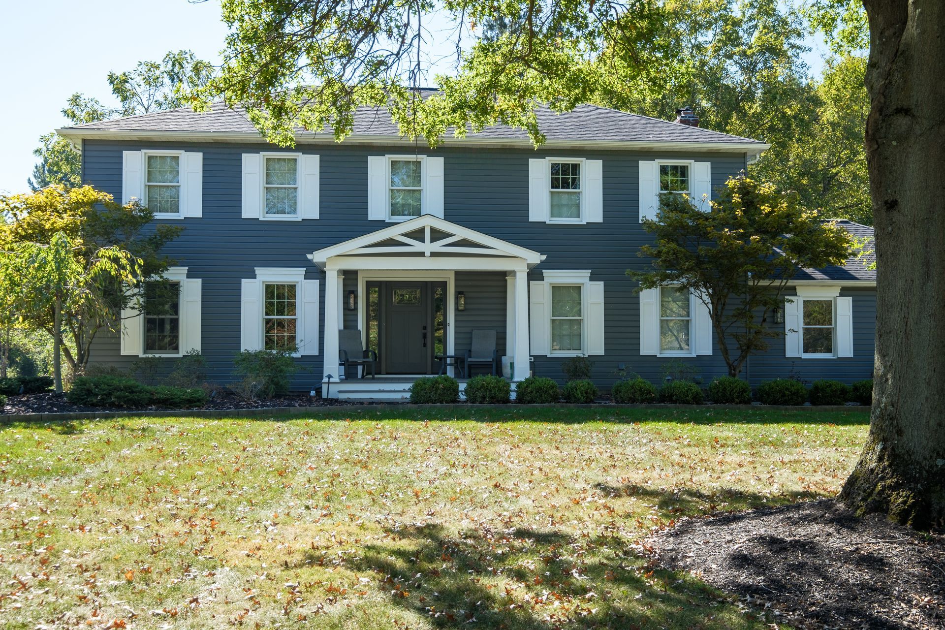 Two-story blue house with white shutters and a covered porch, set on a grassy lawn with a tree in the foreground.
