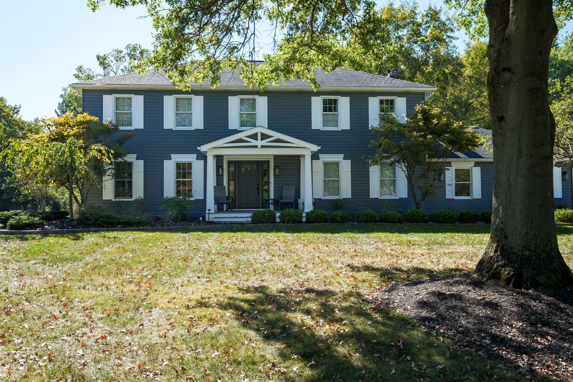 Two-story blue house with white trim and shutters, surrounded by trees, on a sunny day.
