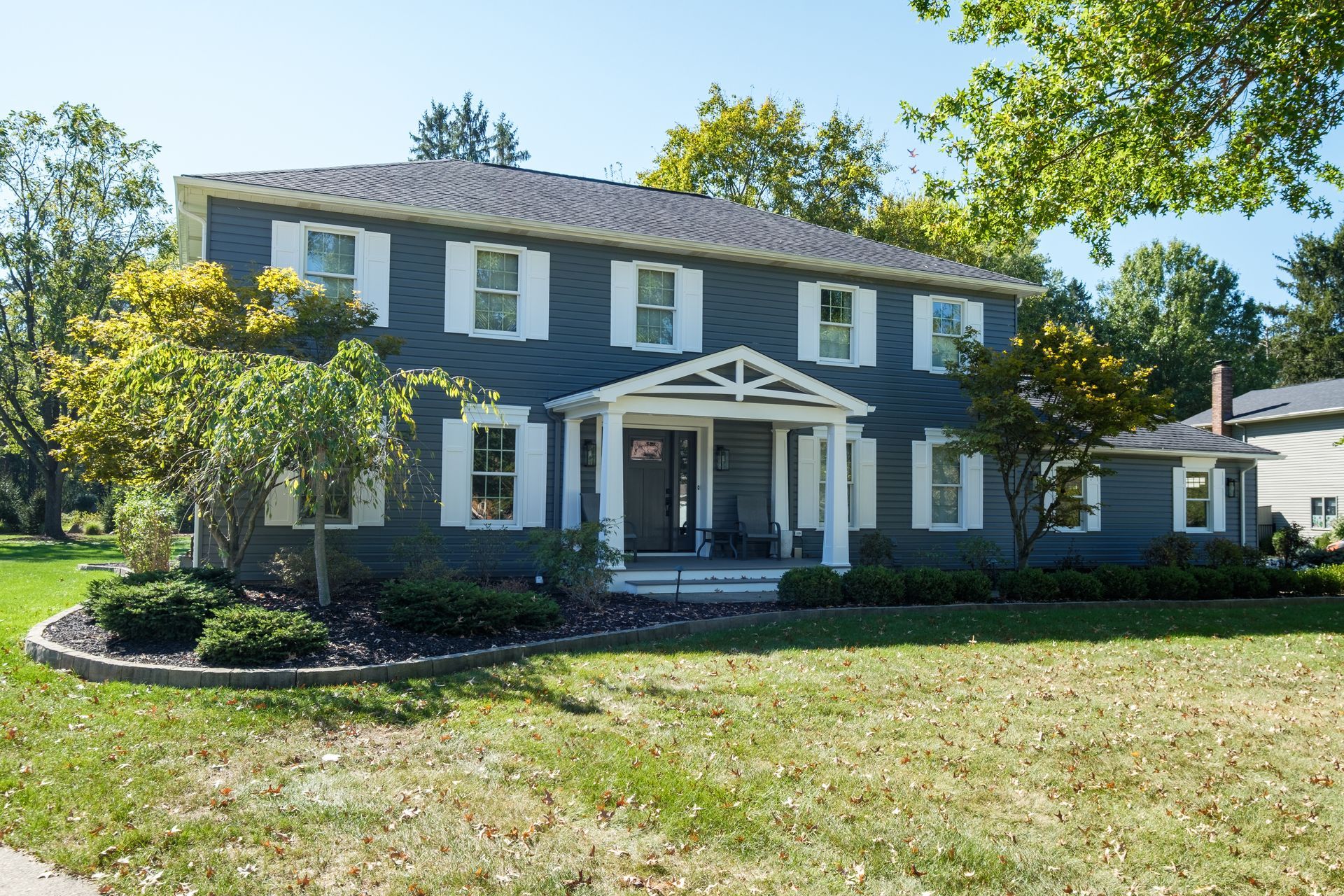 Two-story blue house with white trim and shutters, green lawn, trees, and blue sky.