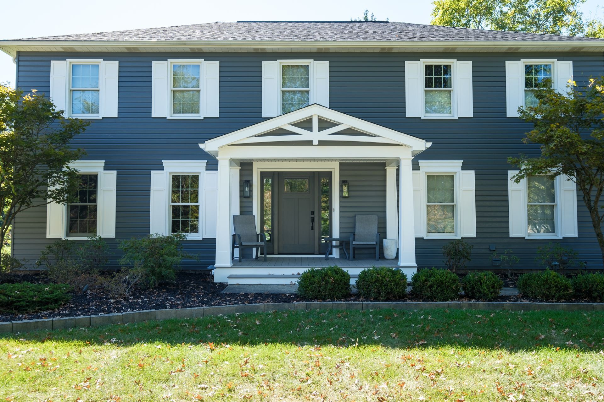 Two-story blue house with white trim, shutters, and a porch. Green lawn and trees in front on a sunny day.