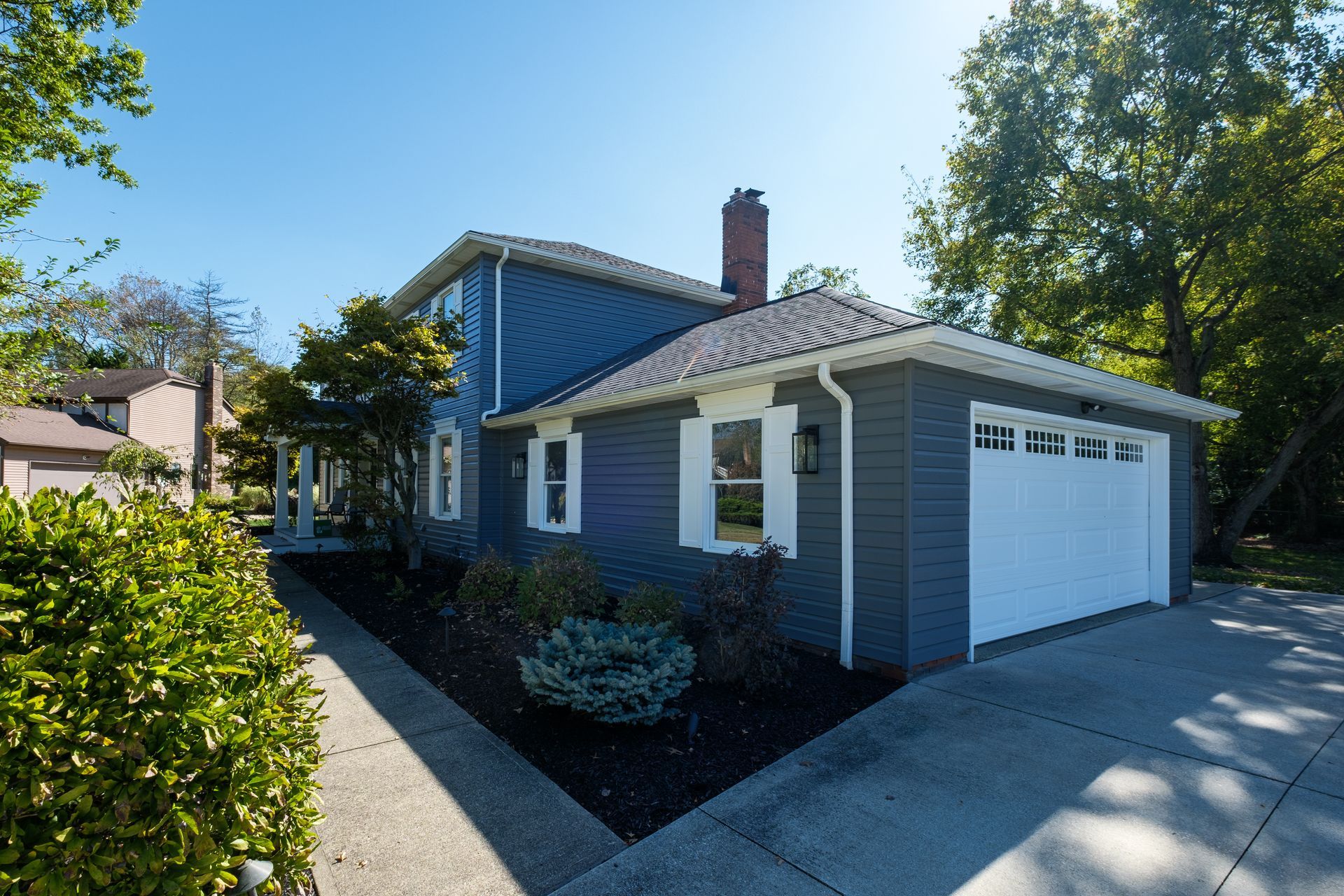 Blue house with white garage doors and shutters, dark trim, and a brick chimney on a sunny day.