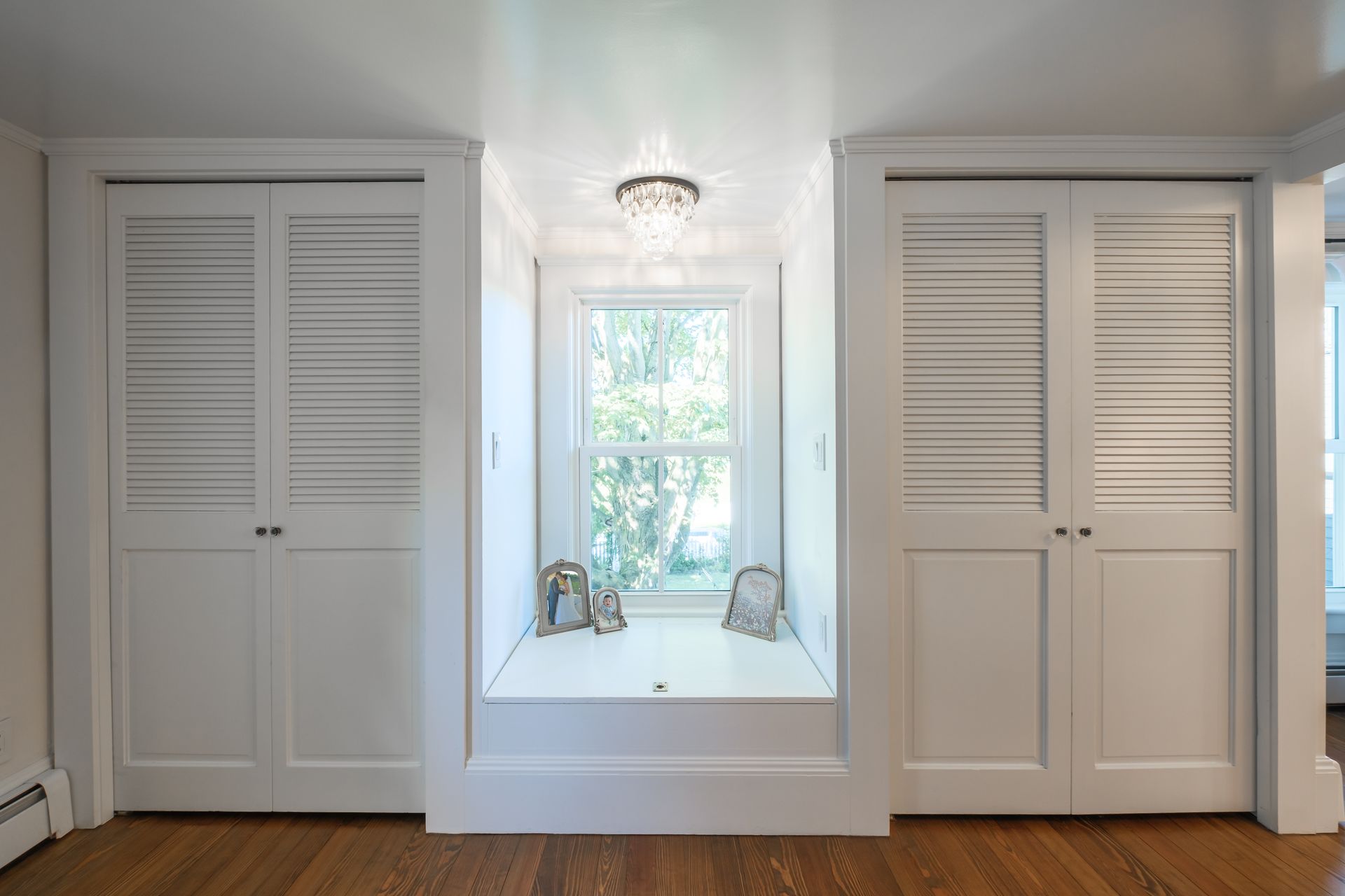 Built-in white closets flank a window seat with a window and decorative frames; overhead chandelier.