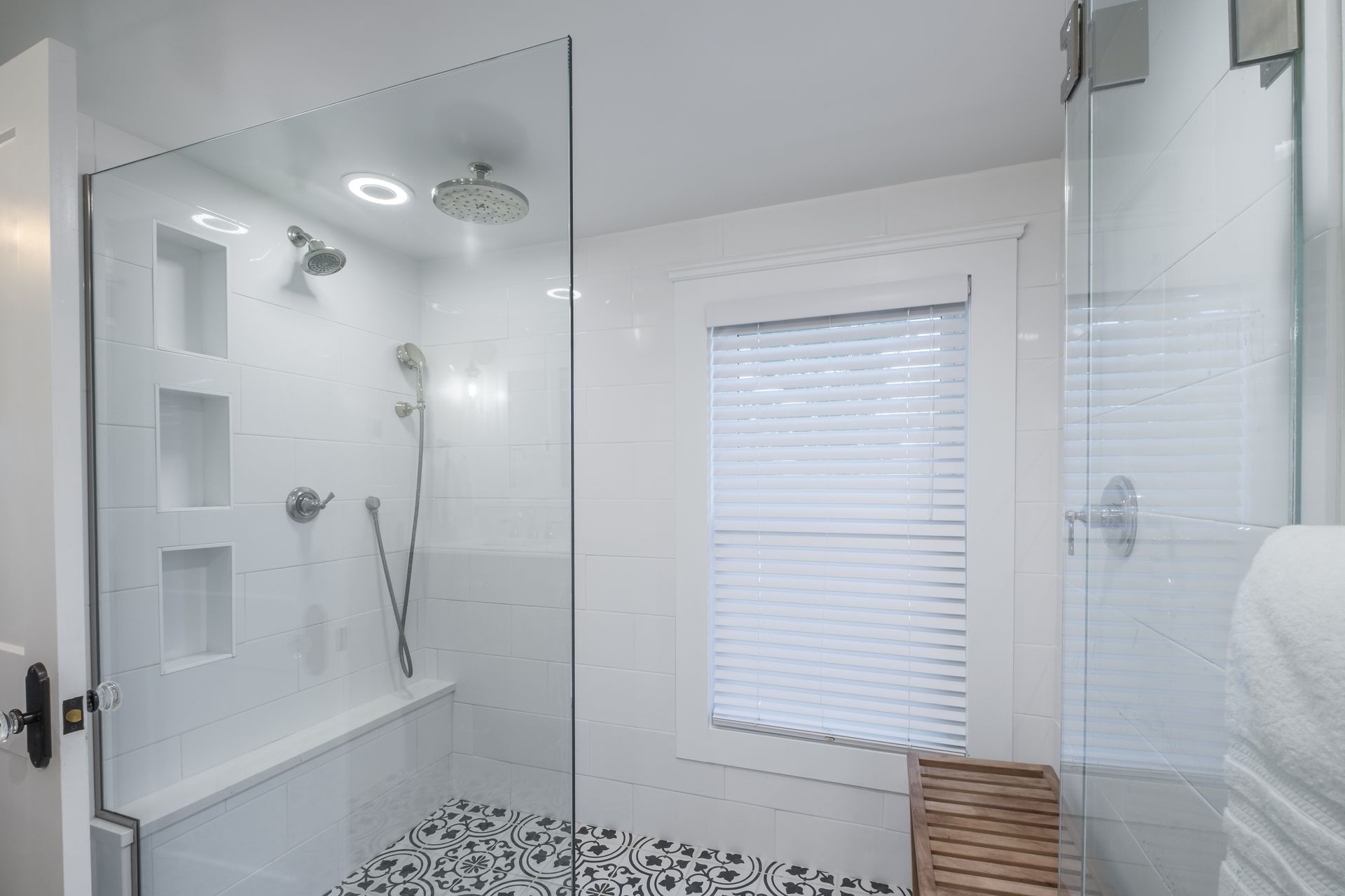 White tiled shower with glass door, pebble floor, built-in bench, and window with closed blinds.
