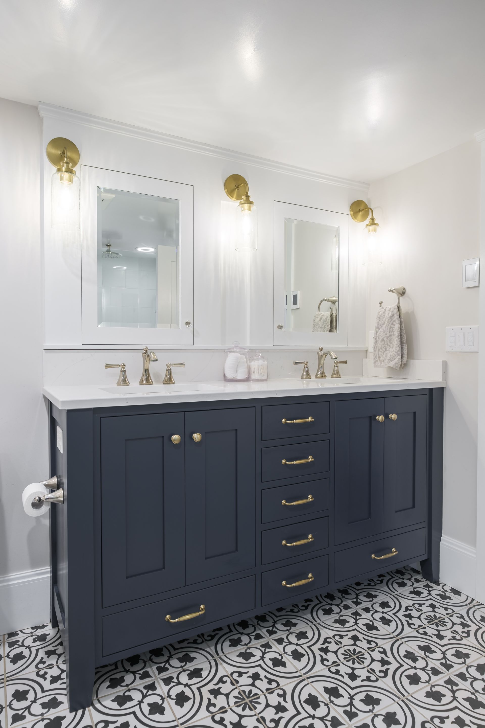 Navy blue double bathroom vanity with gold hardware, white countertop, patterned floor, and gold sconces.
