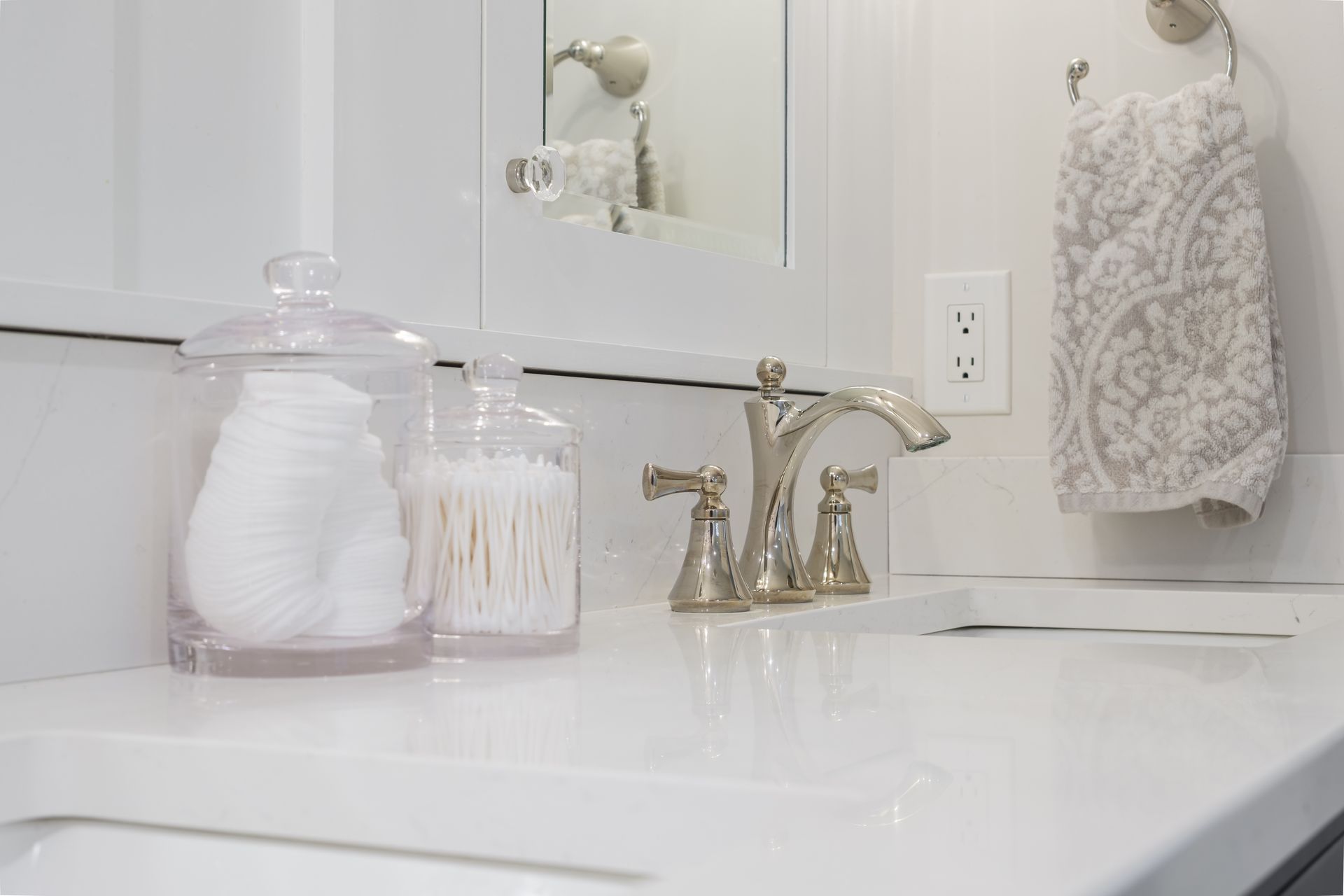 Bathroom countertop with cotton balls, Q-tips, and a faucet. Towel on a hook. White and silver tones.