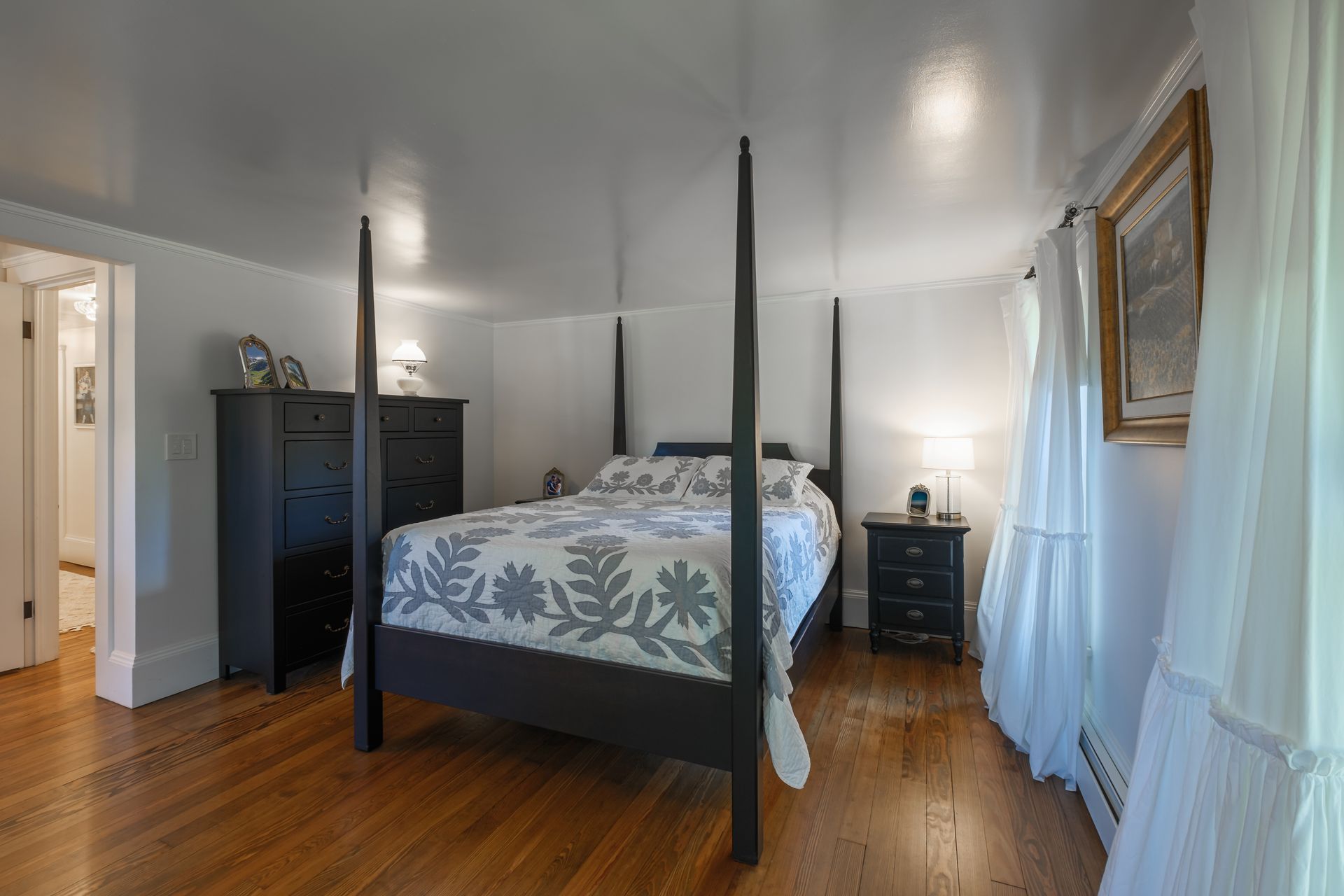 Bedroom with a dark wood four-poster bed, black dresser, and nightstand on a hardwood floor. White walls.