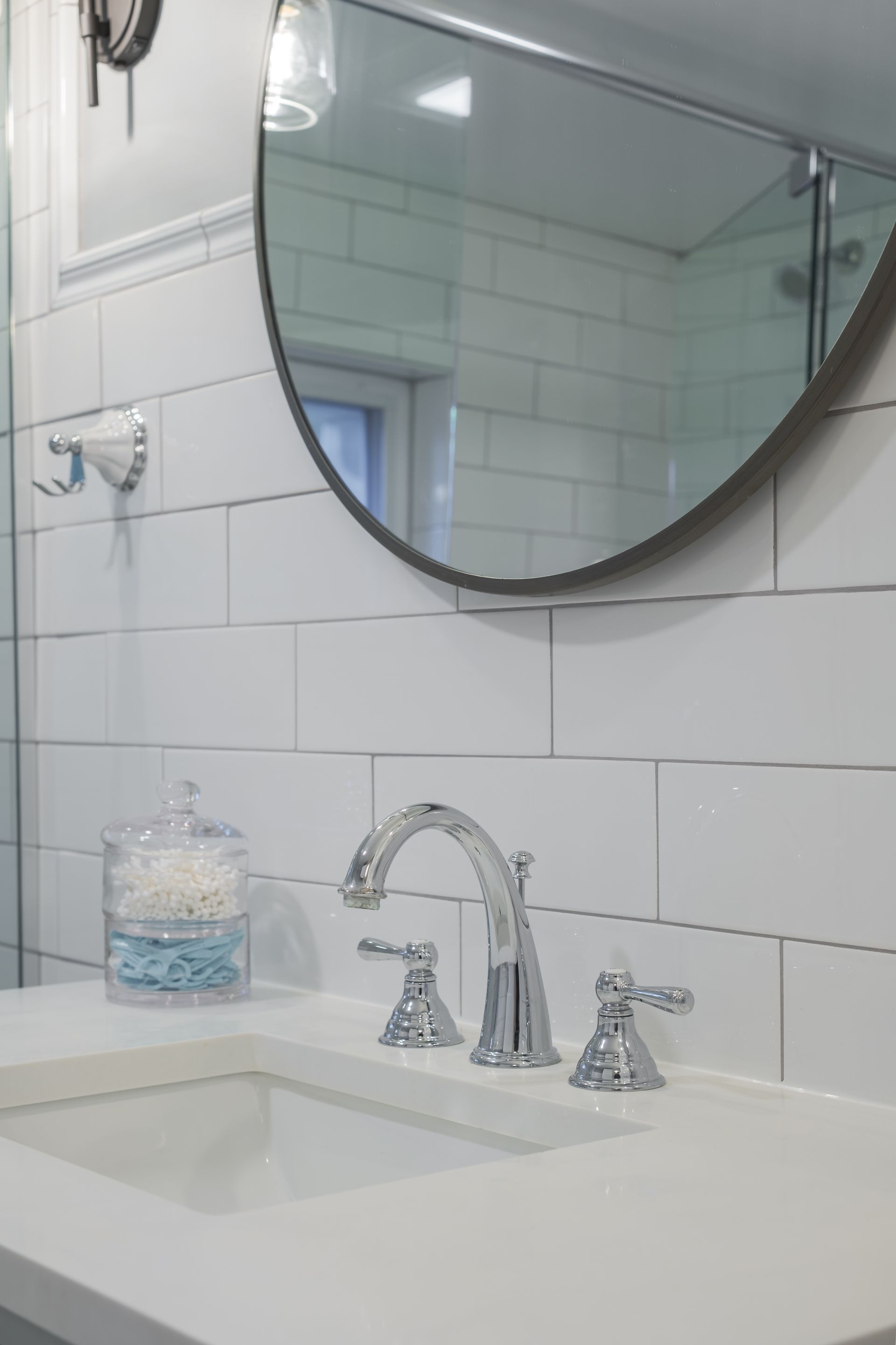 Bathroom vanity with chrome faucet, oval mirror, white subway tile, and a cotton swab jar.