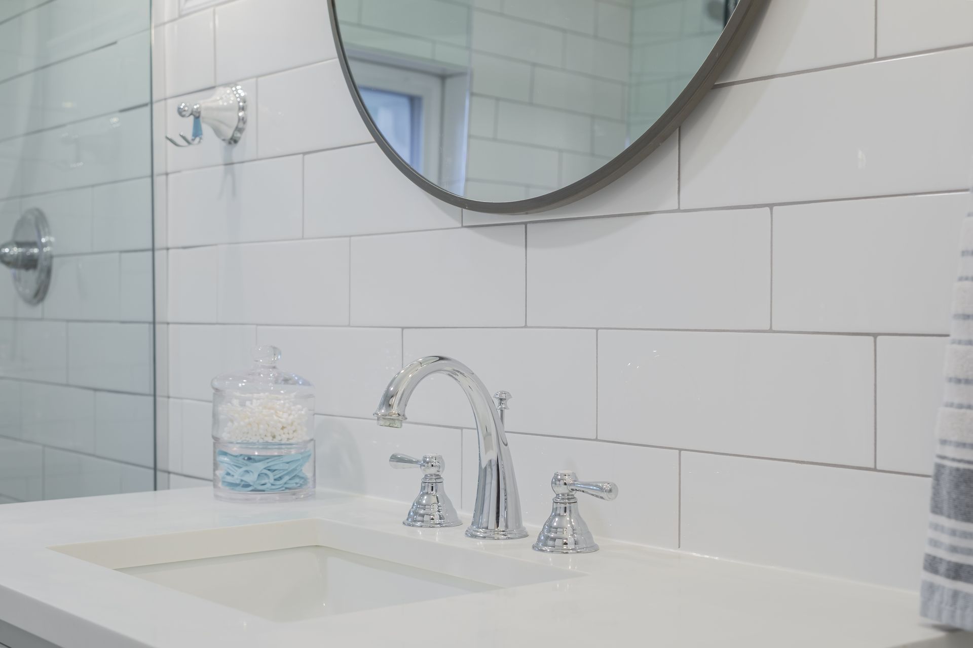 Bathroom sink with chrome faucet, white countertop, white subway tile wall, round mirror.