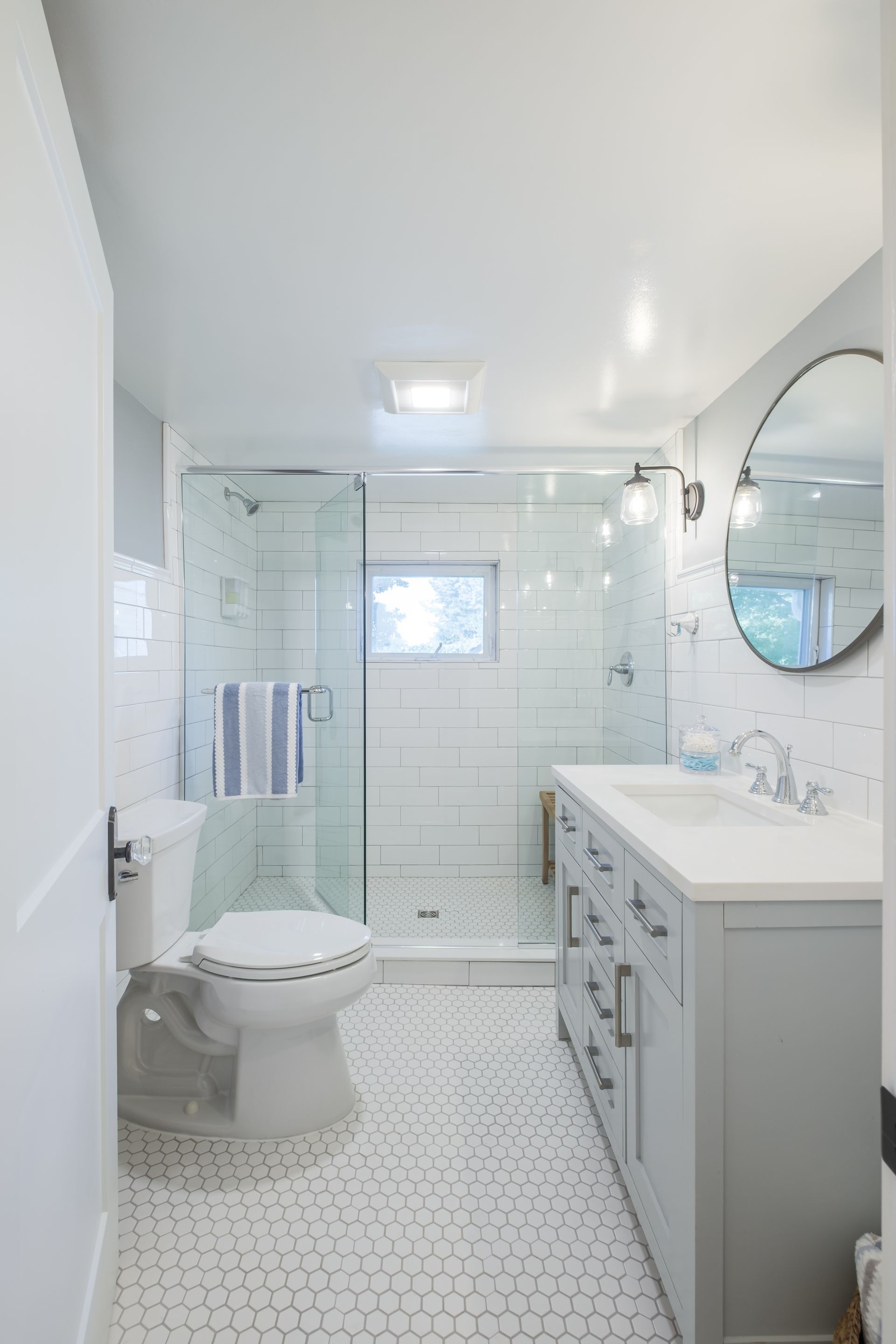 Bathroom with white and gray tones, glass shower, vanity, toilet, and round mirror.