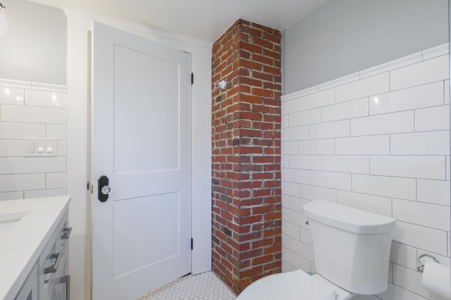Bathroom with white tile, a white toilet, exposed brick column, and a white door.
