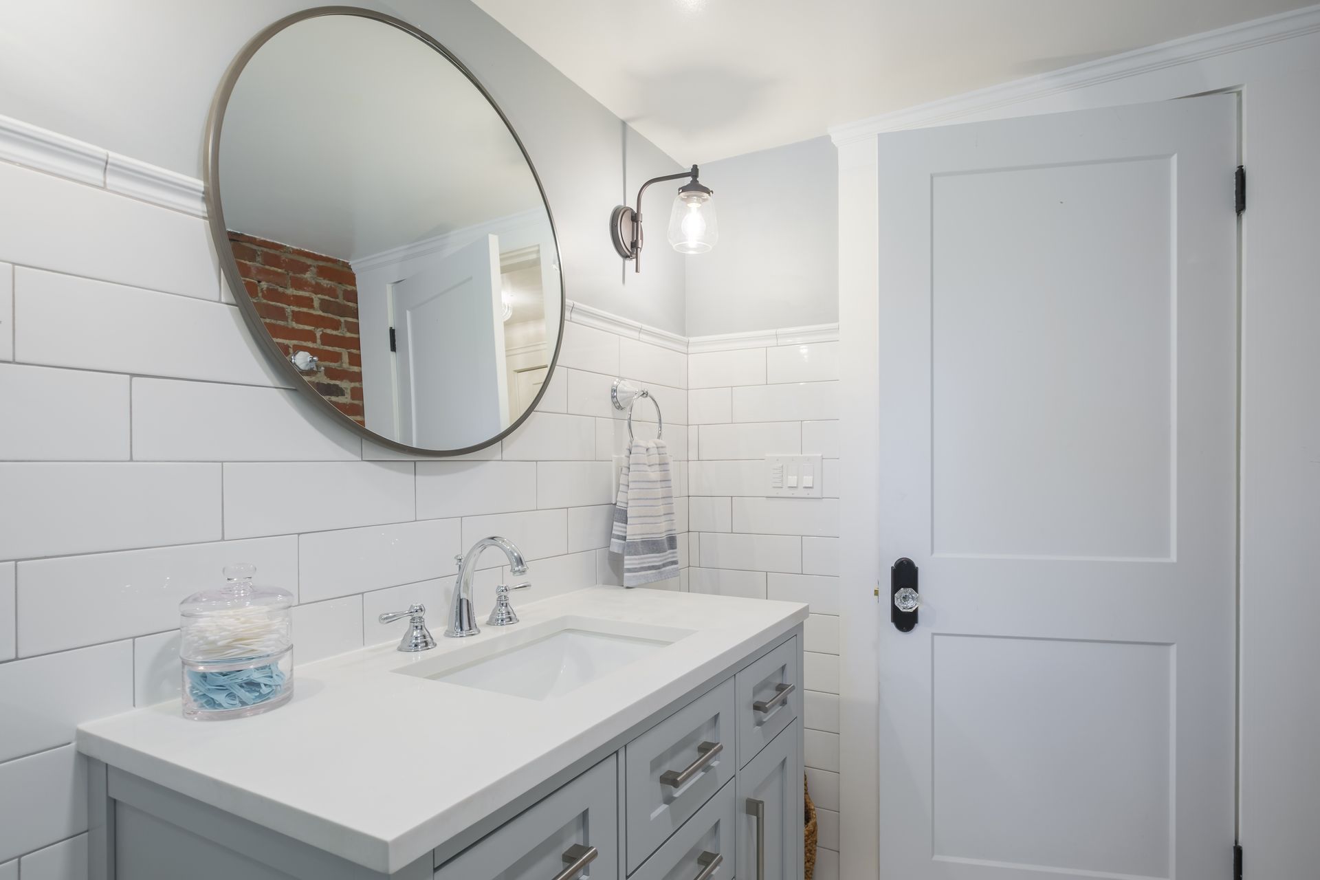 Bathroom with white subway tile, grey vanity, round mirror, and a white door.