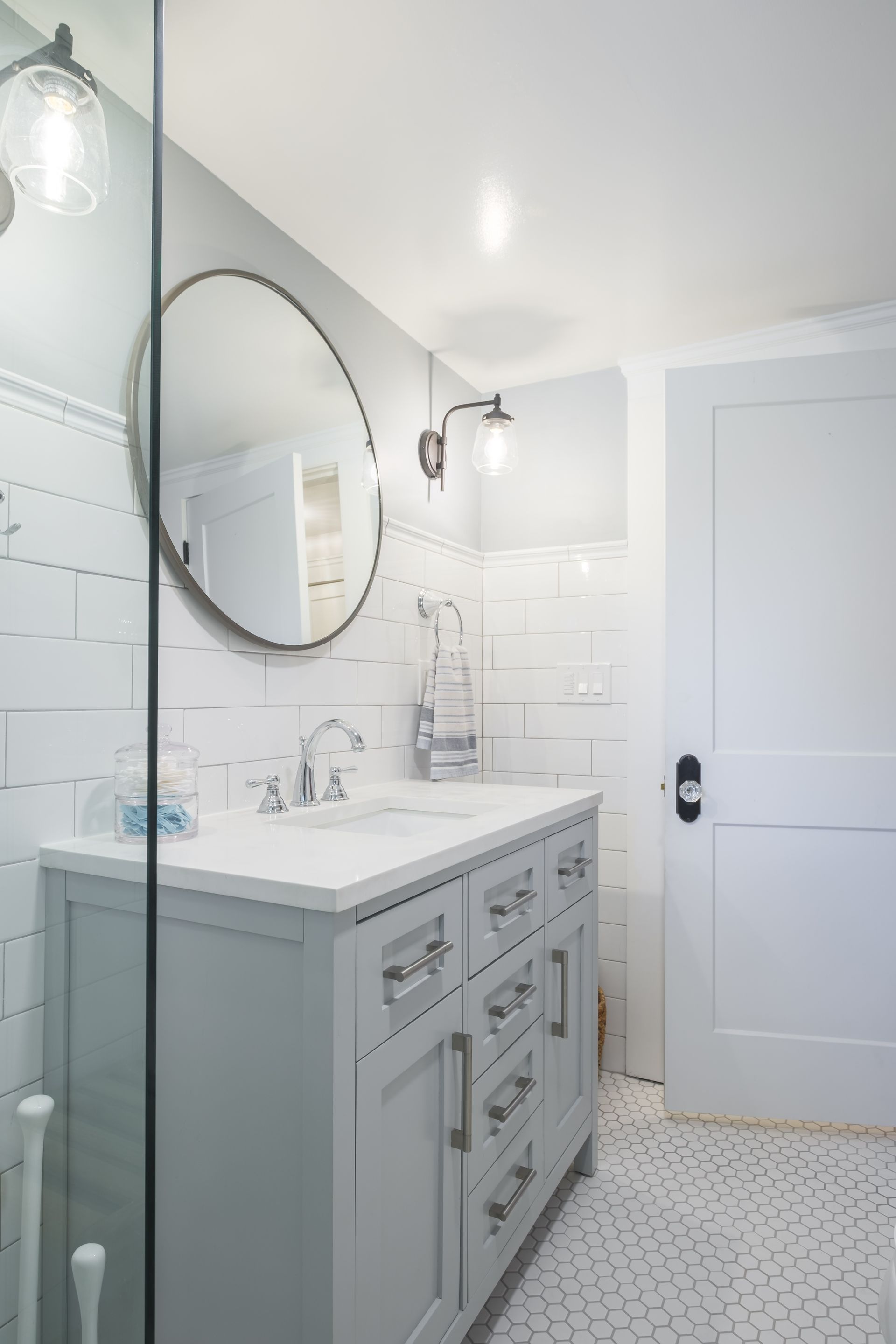 Bathroom with gray vanity, round mirror, white subway tile, and a light-gray door.
