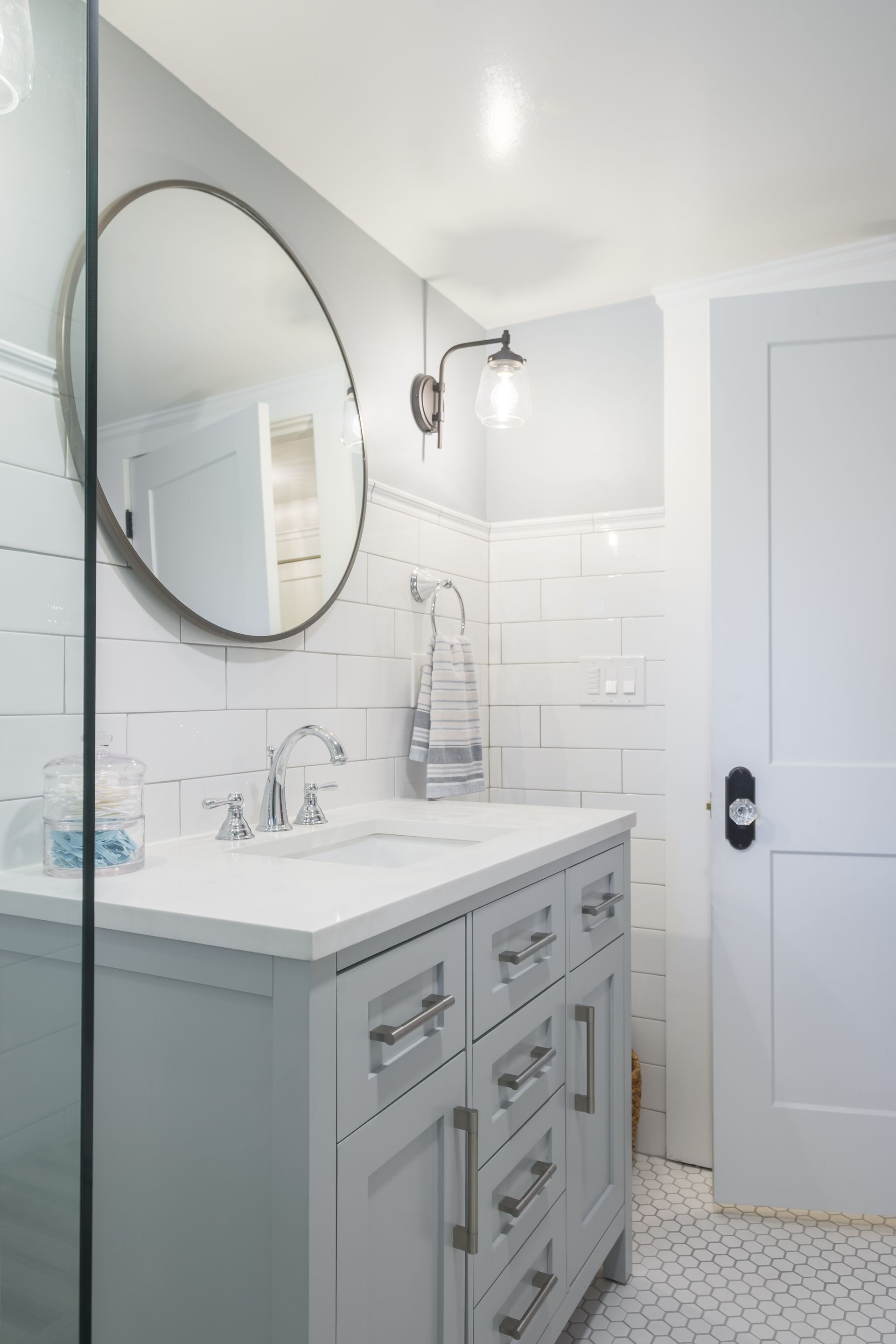 A light gray bathroom with a white vanity, round mirror, and white tiled walls.