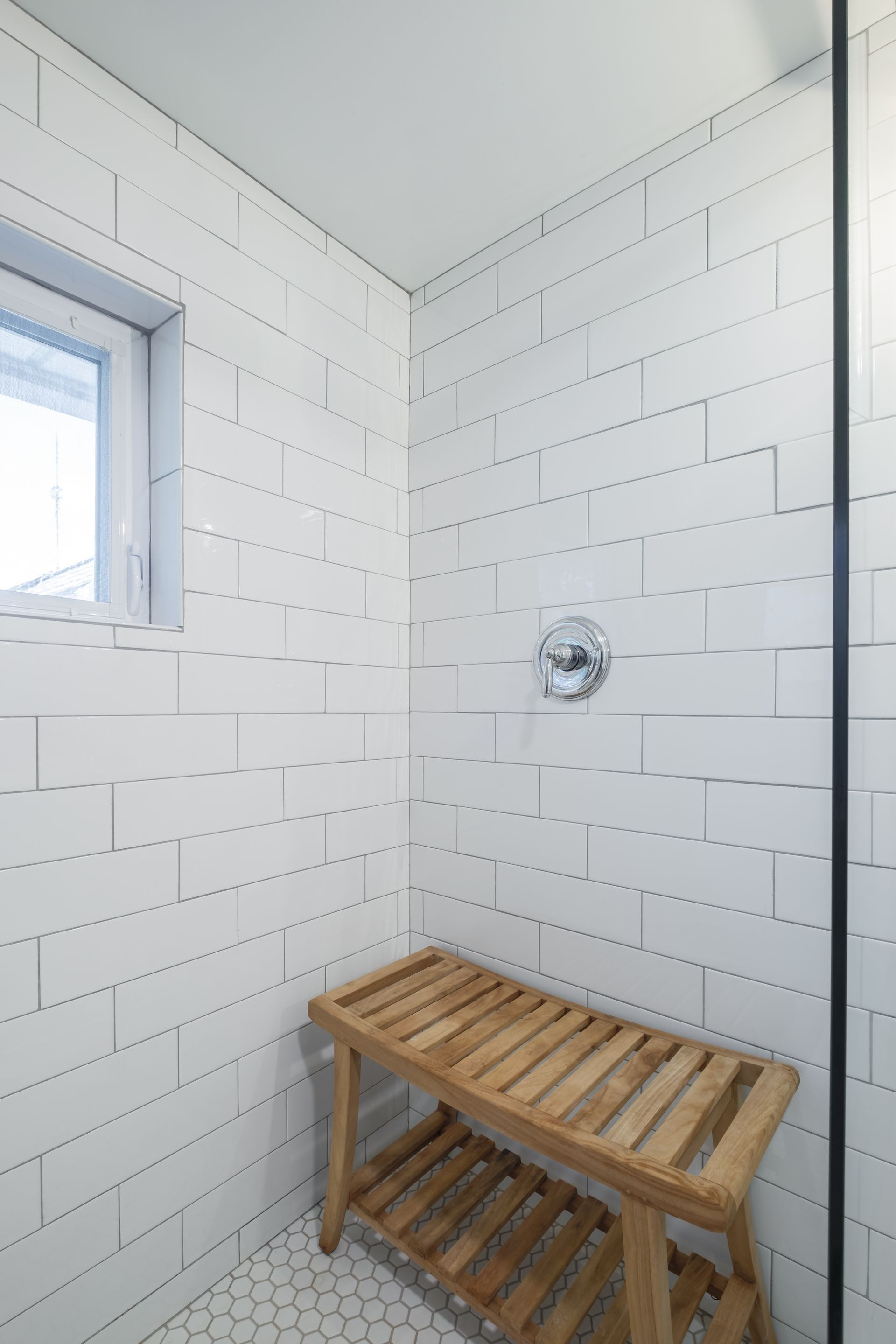 A white-tiled shower with a wooden bench, window, and silver shower controls.