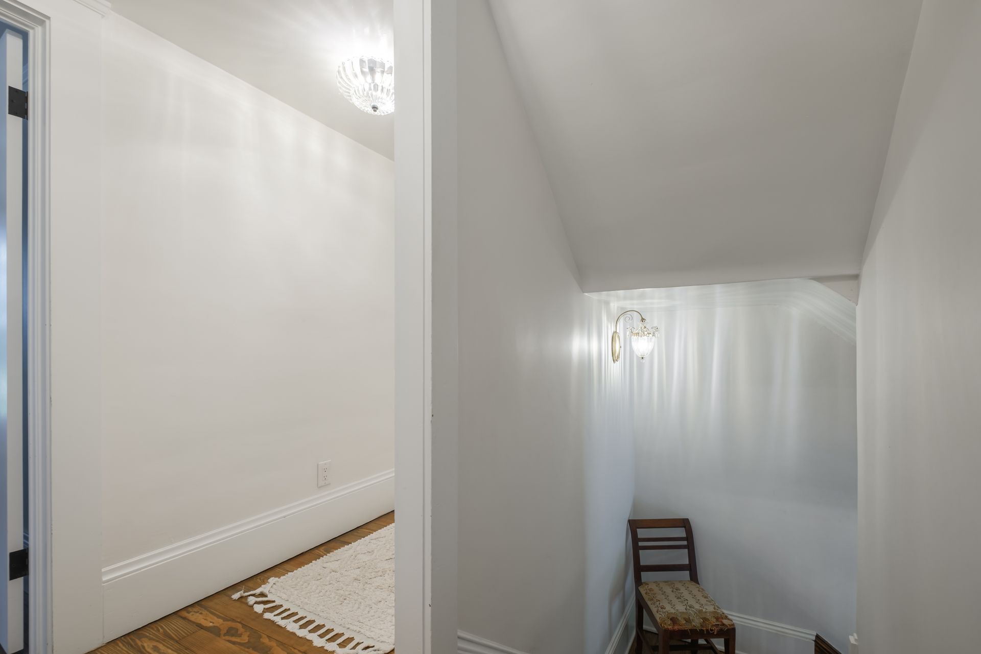 Hallway with white walls, a chair, and a glimpse into a room with a chandelier.