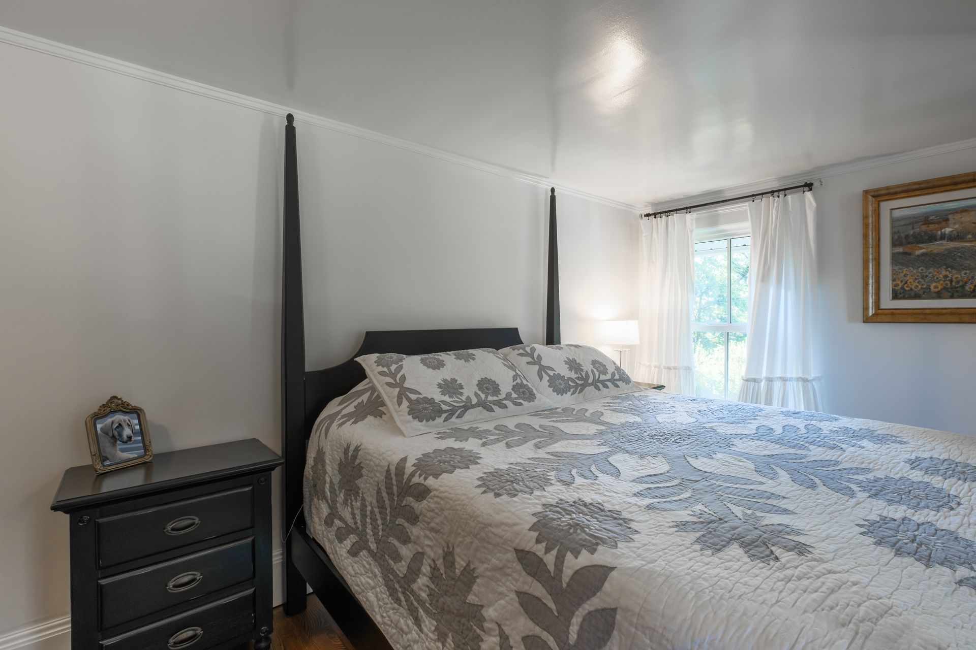 Bedroom with a bed, nightstand, window with curtains, and artwork. The room is painted white.