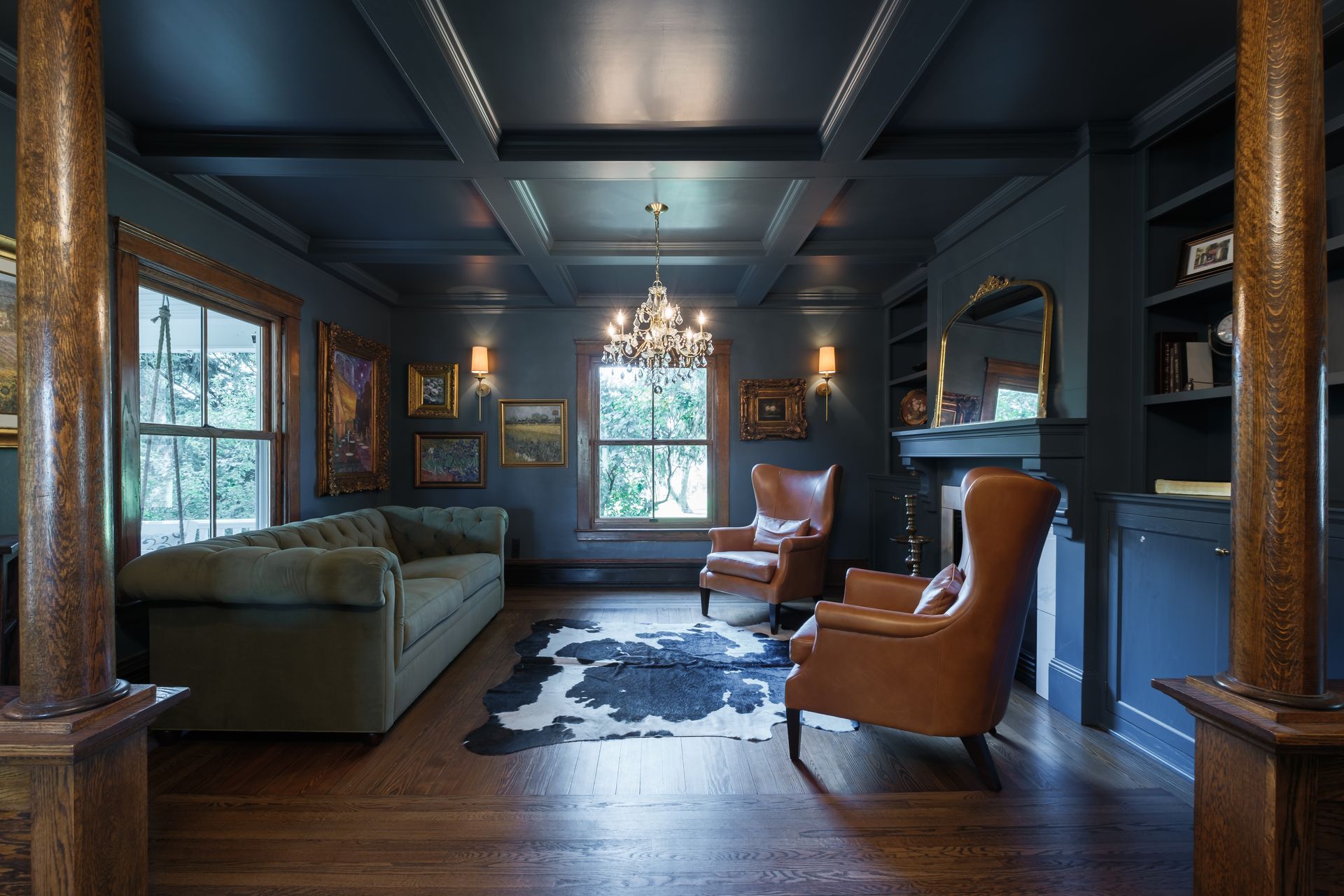 Dark blue-painted room with a green couch, leather chairs, and chandelier. Wooden floor, rug, and windows.