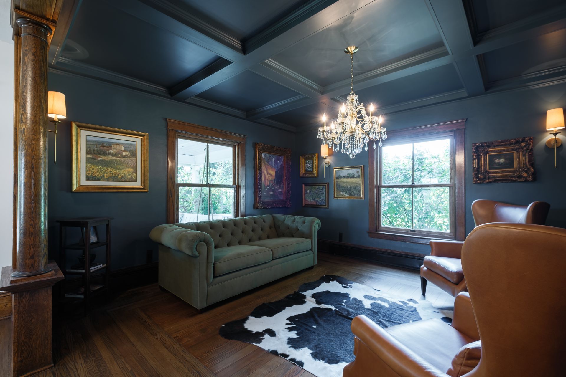 Dark room with green sofa, two leather chairs, chandelier, art, cowhide rug, wood floors, and windows.