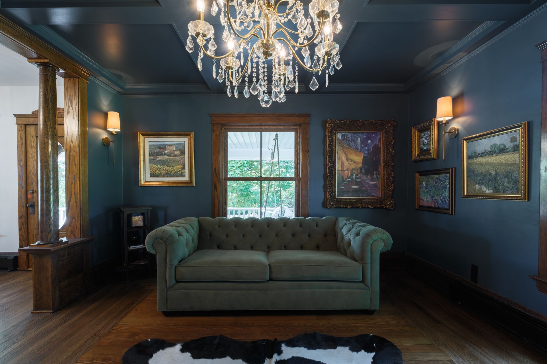 Dark blue room with green couch, paintings, chandelier, wood floor, and a cowhide rug.