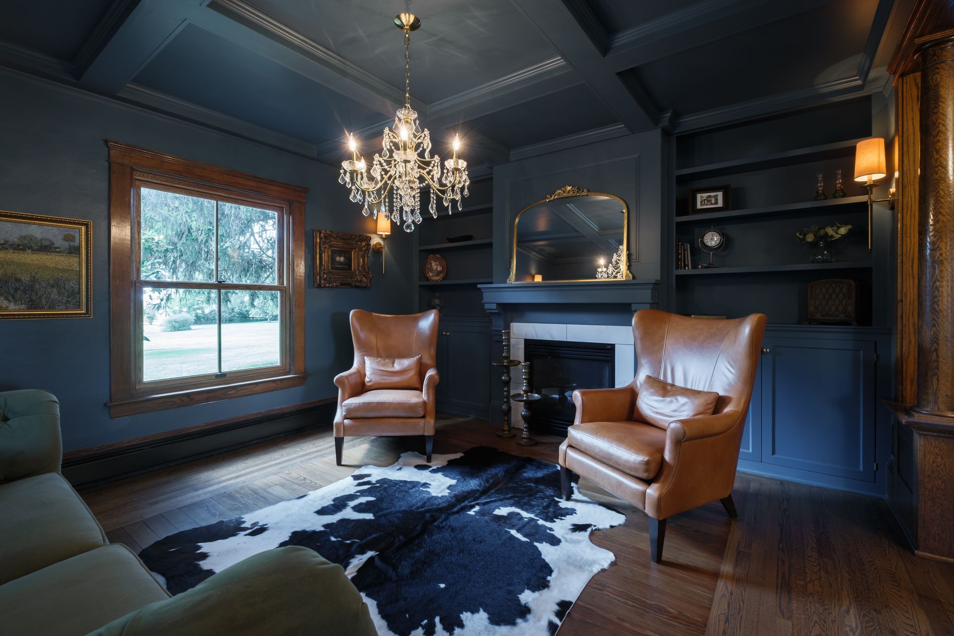 Cozy study with two leather armchairs, a cowhide rug, fireplace, and chandelier; dark blue walls and wood trim.
