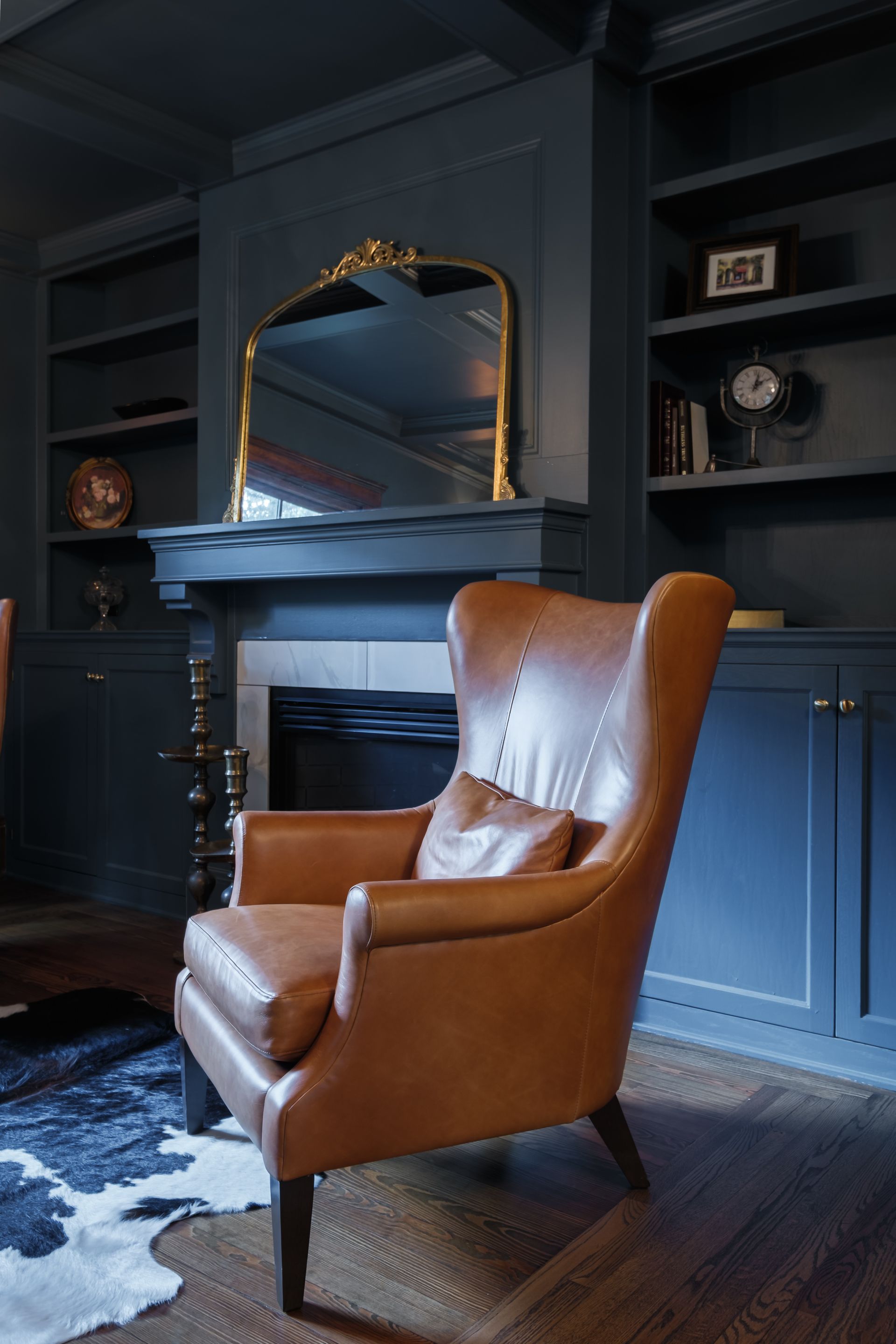 Leather armchair in a dark blue room, gold-framed mirror over fireplace, built-in bookshelves.