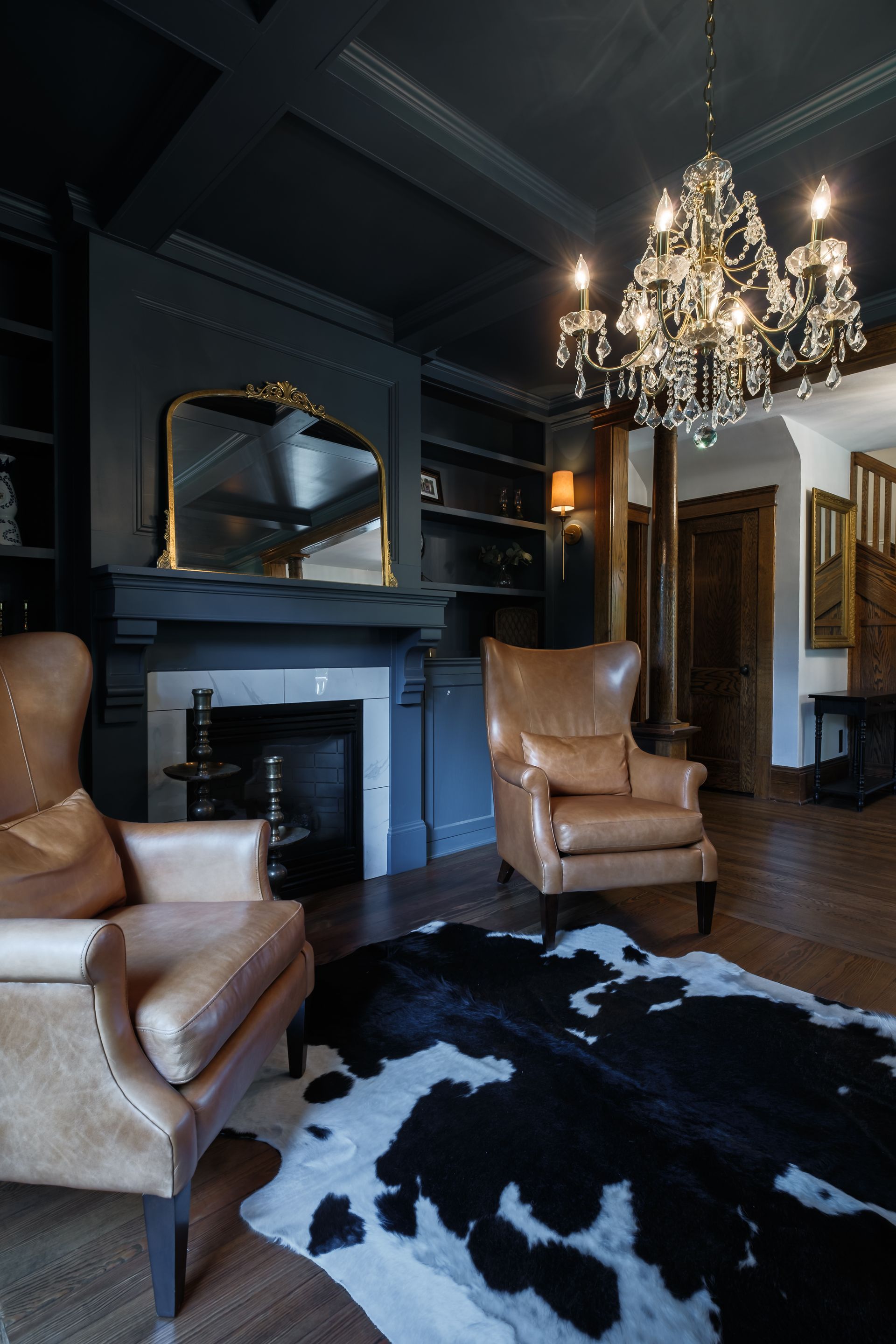 Cozy library with dark blue walls, leather chairs, a cowhide rug, fireplace, and chandelier.