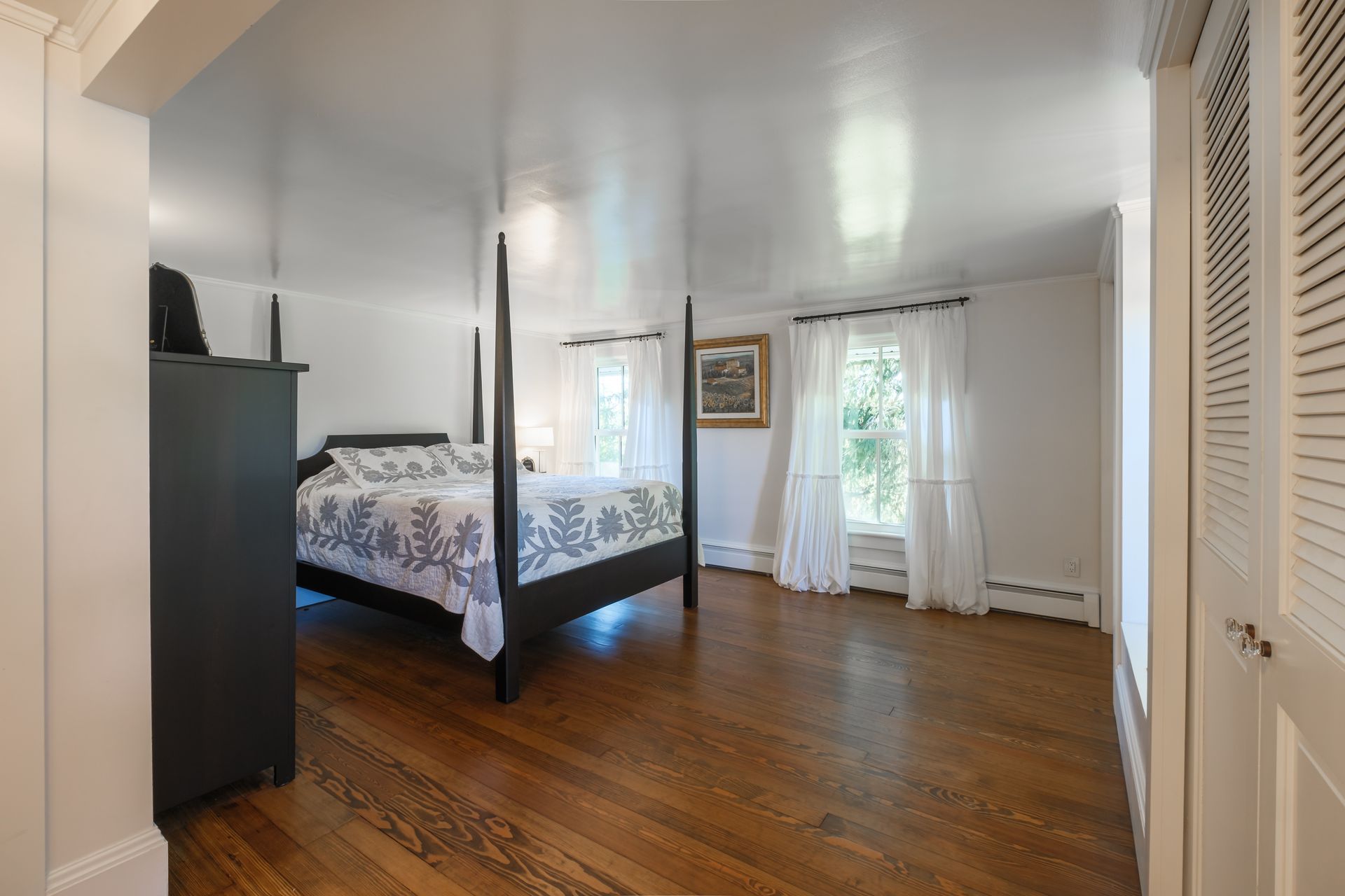 Bedroom with dark wood floor, four-poster bed, white curtains, and a dark dresser.