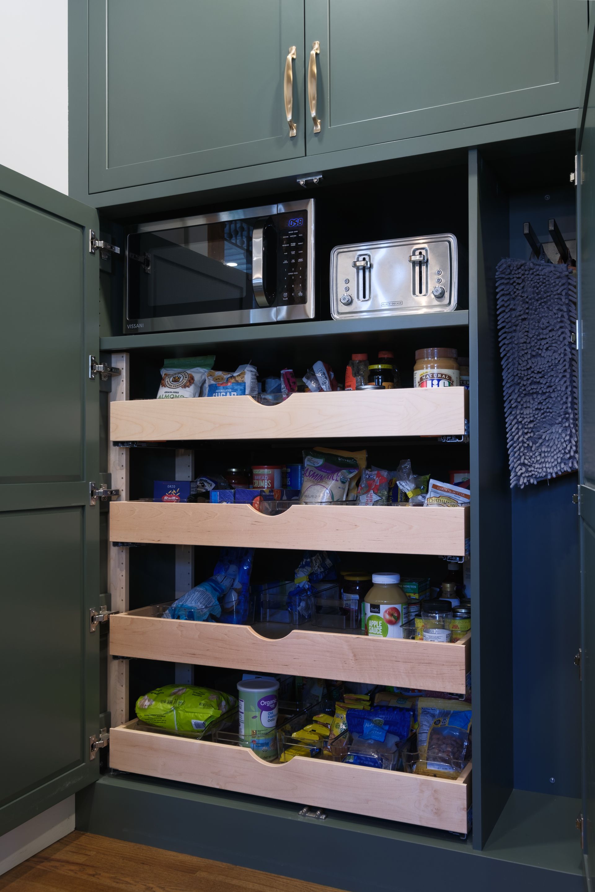 Green kitchen cabinet with microwave, toaster, and pull-out shelves filled with food.