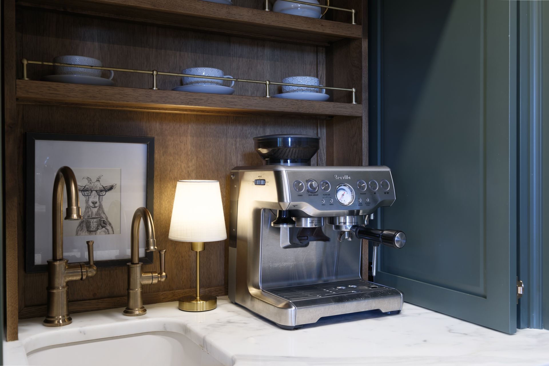 Espresso machine on counter with lamp, art, faucet, and shelf holding cups.