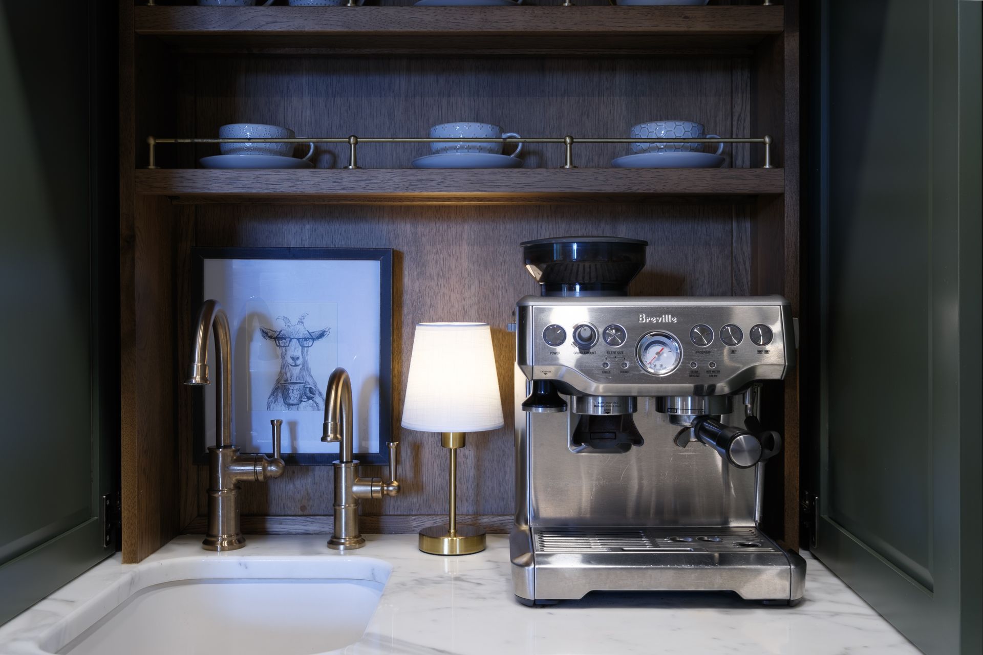 Espresso machine, sink, and lamp in a built-in coffee station with cups on a shelf.