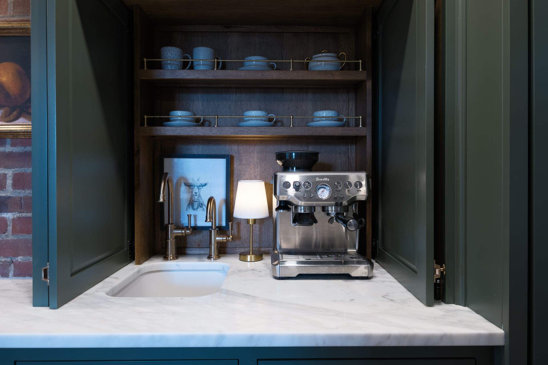 Built-in coffee station with an espresso machine, sink, and shelves holding cups; forest green cabinets, marble countertop.