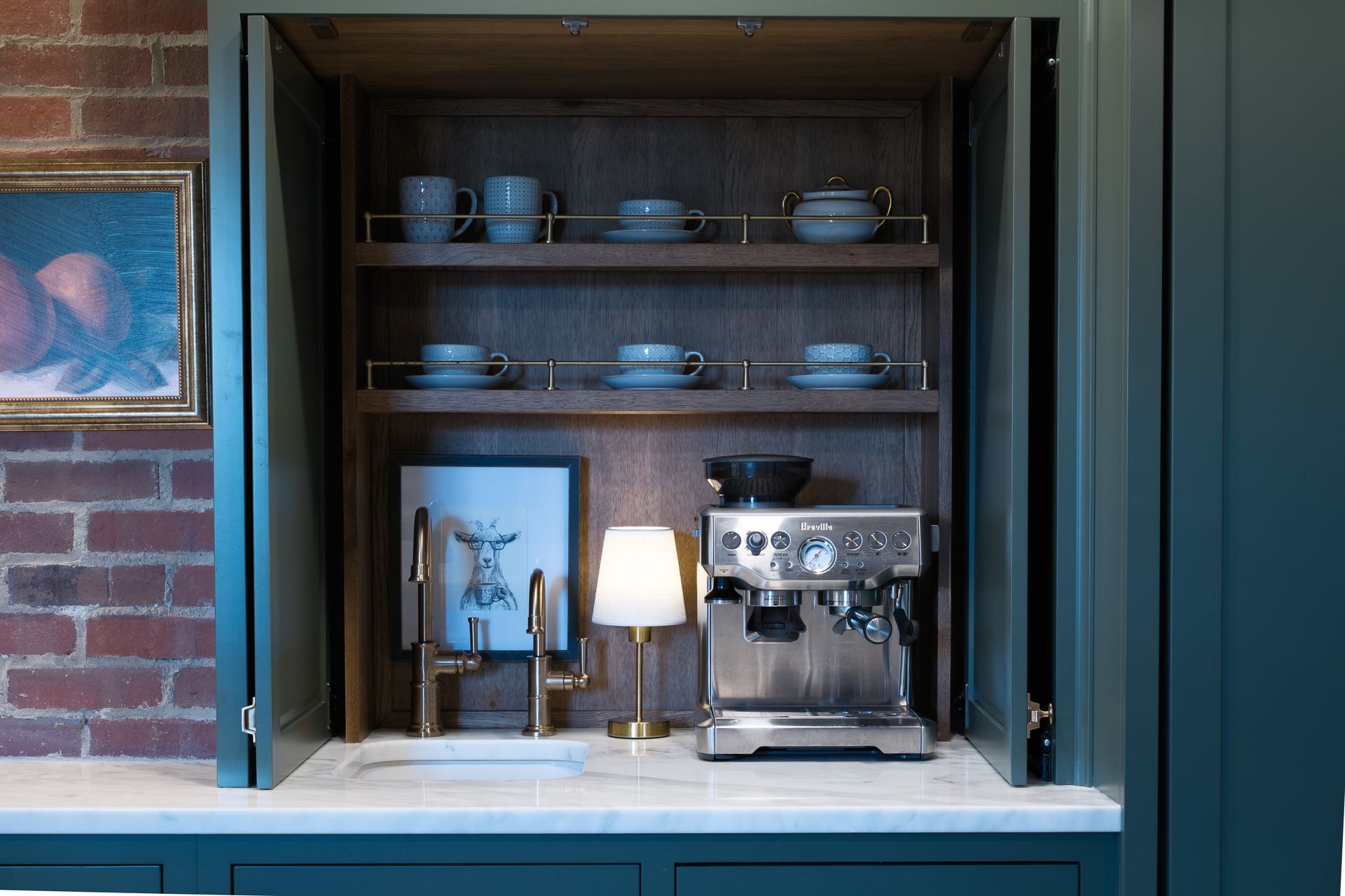 Coffee station built into dark green cabinets with brick wall, lit by small lamp.