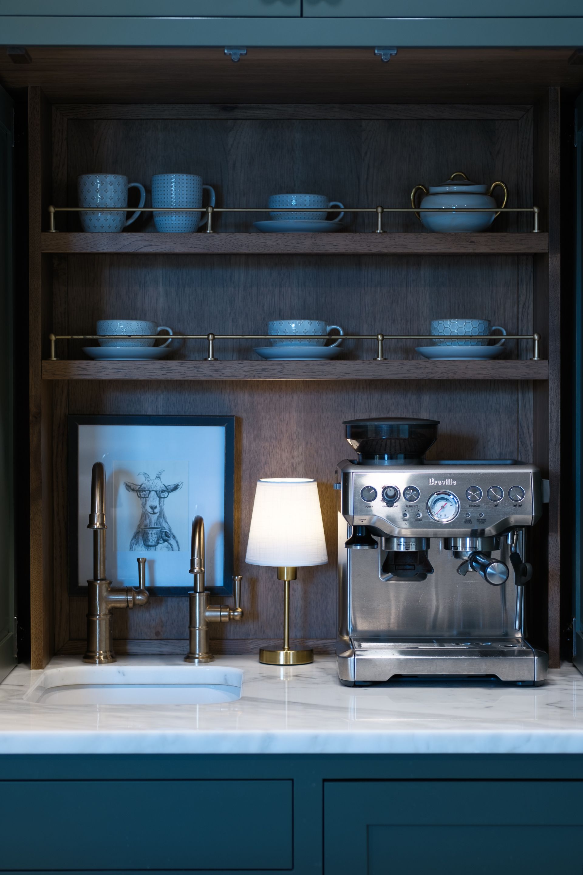 Coffee station with espresso machine, mugs on shelves, sink, lamp, and artwork, set in a dark wood cabinet.