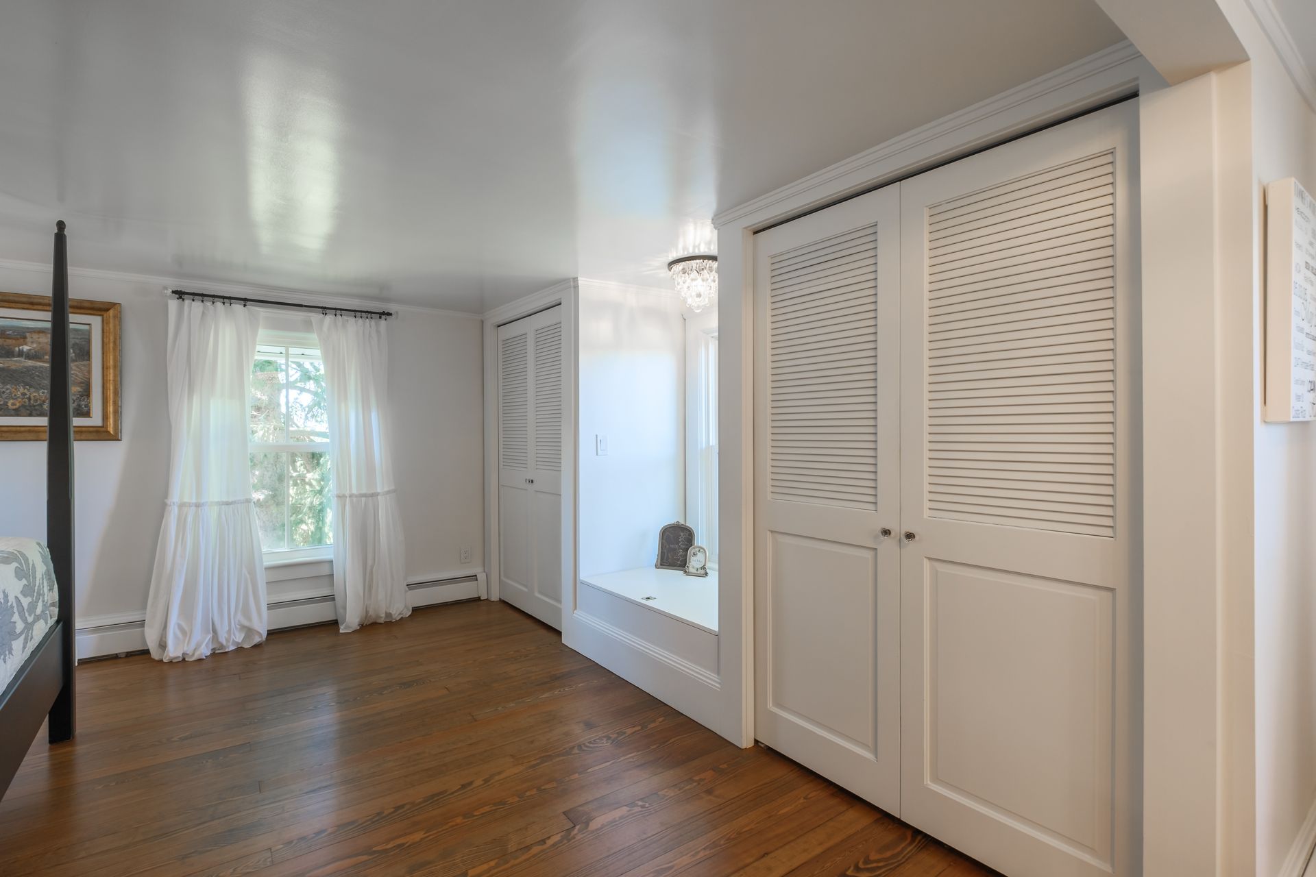 Bedroom interior with hardwood floors, white walls and closets, window with sheer curtains, and a four-poster bed frame visible.