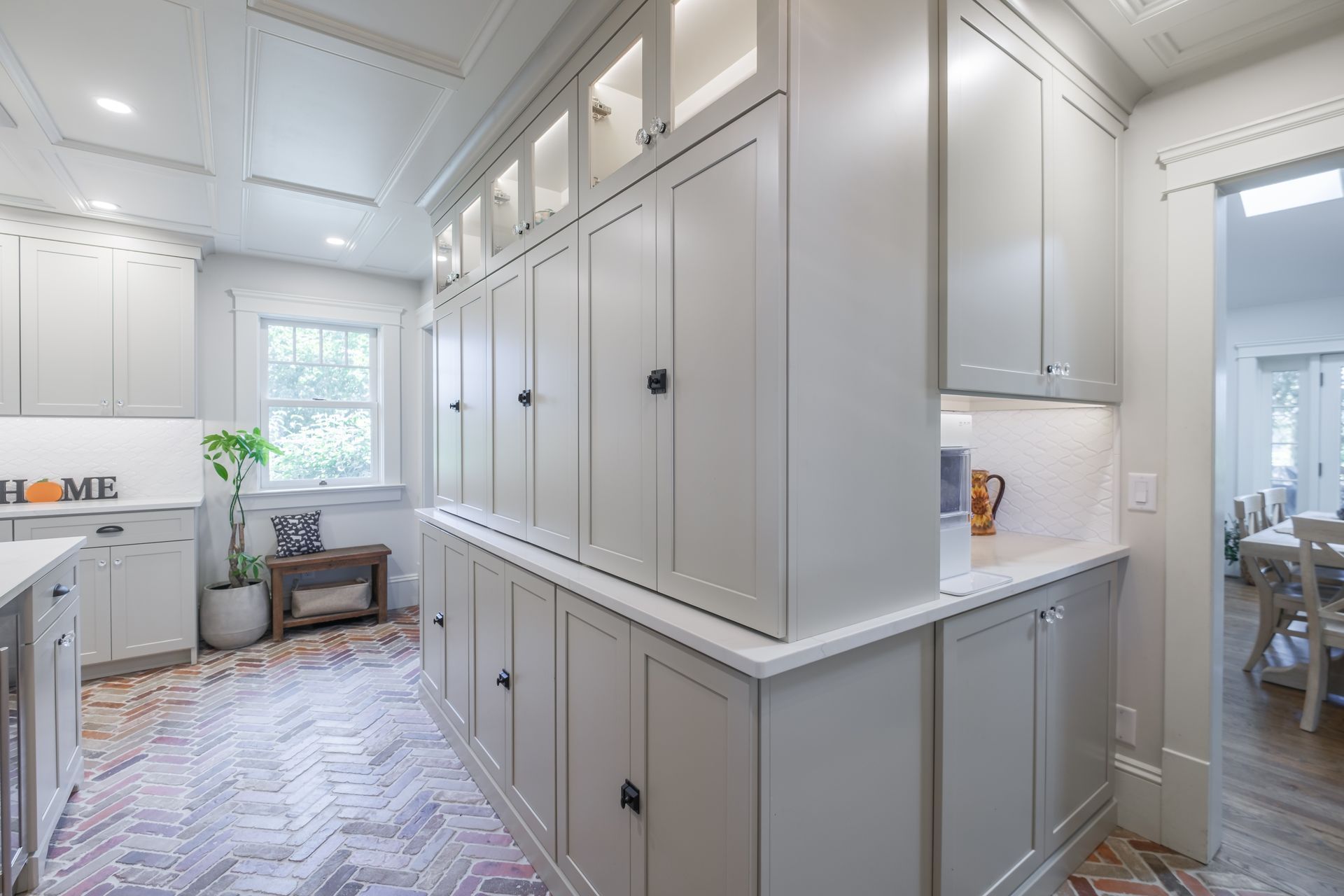 A bright kitchen with light gray cabinetry, brick floor, and an open doorway to a dining room.