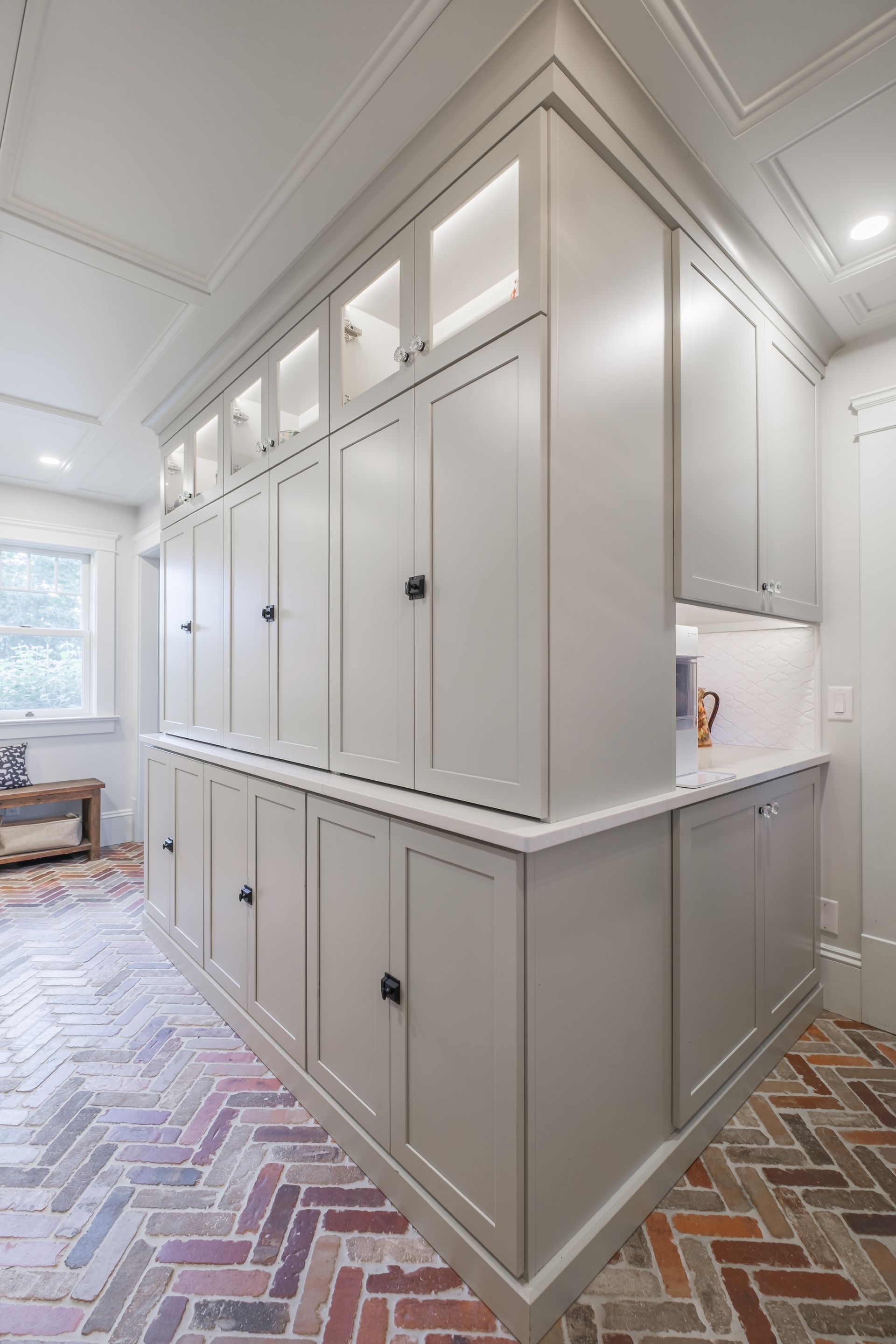 Light gray cabinets in a corner with overhead lighting, white countertop, and herringbone brick floor.
