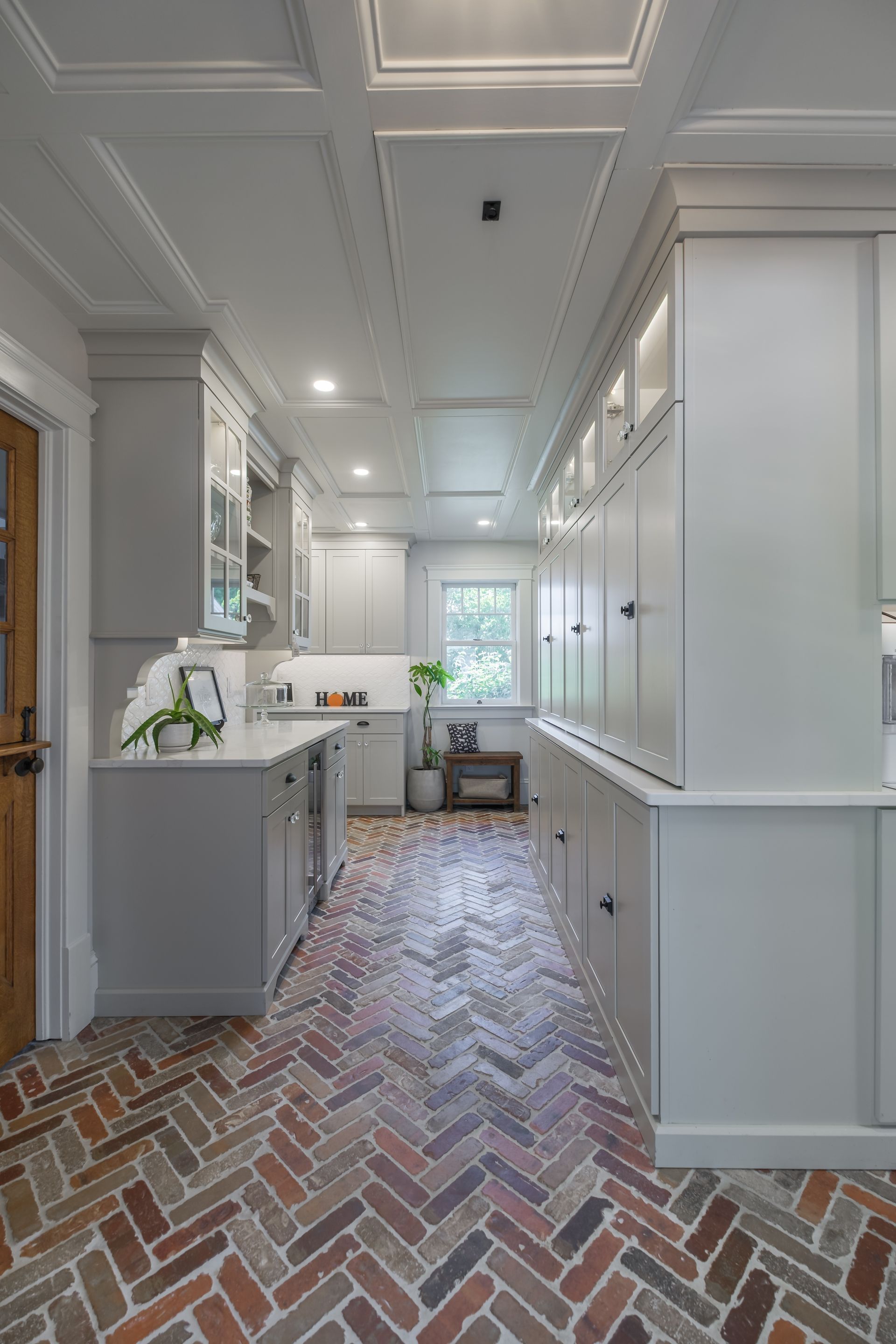 Narrow kitchen with herringbone brick floor, gray cabinets, and coffered ceiling.