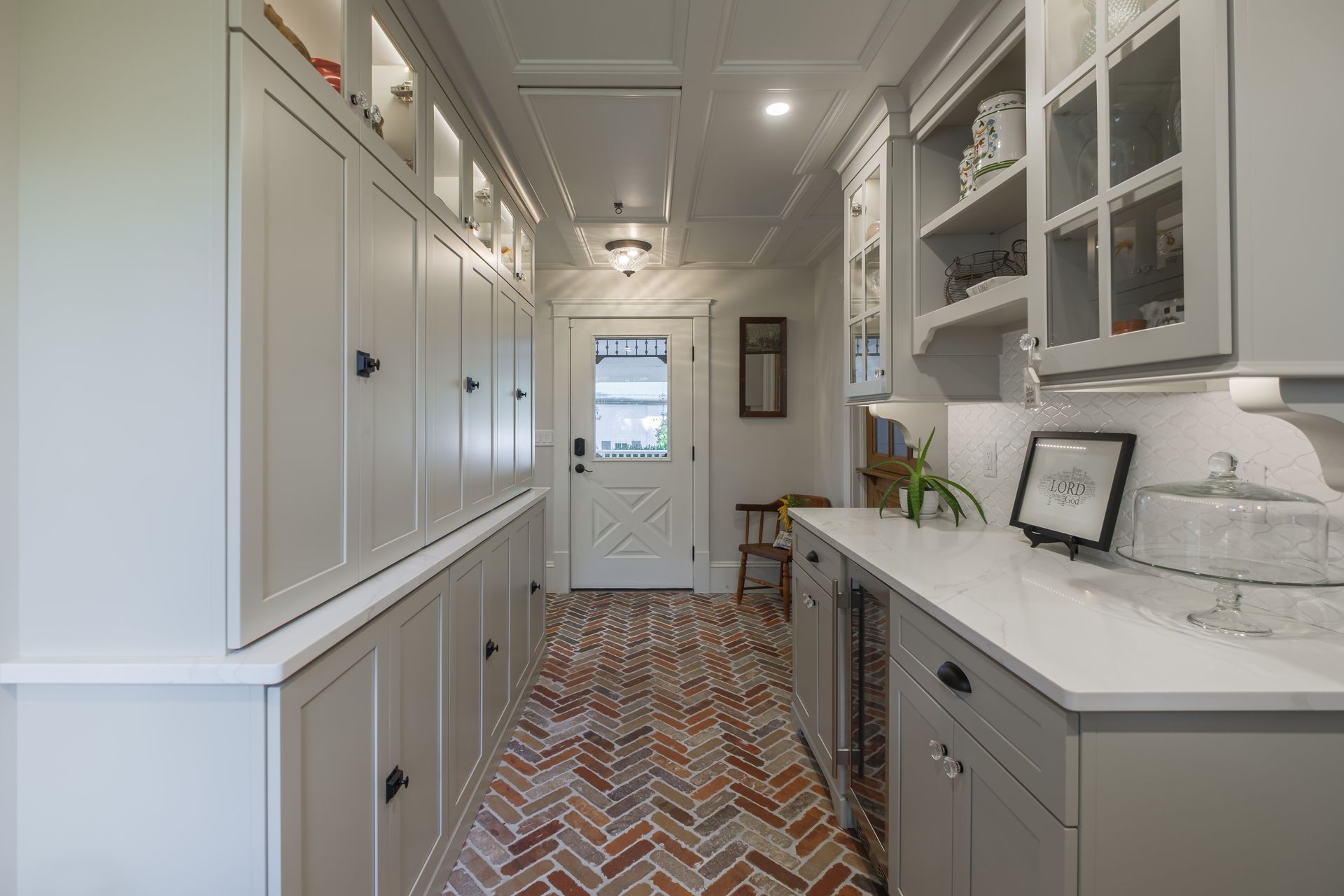 Narrow pantry with gray cabinets, white countertops, brick floor, and a door at the end.