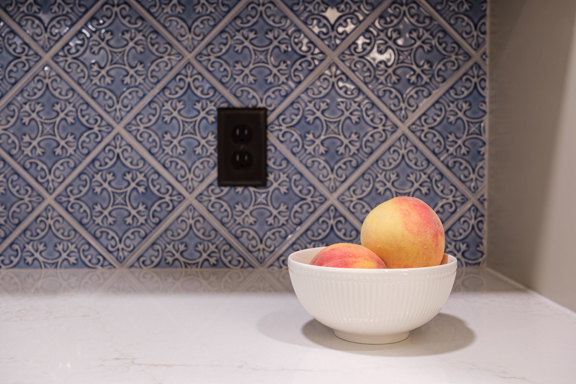 A bowl of peaches is sitting on a counter in front of a blue tile wall.