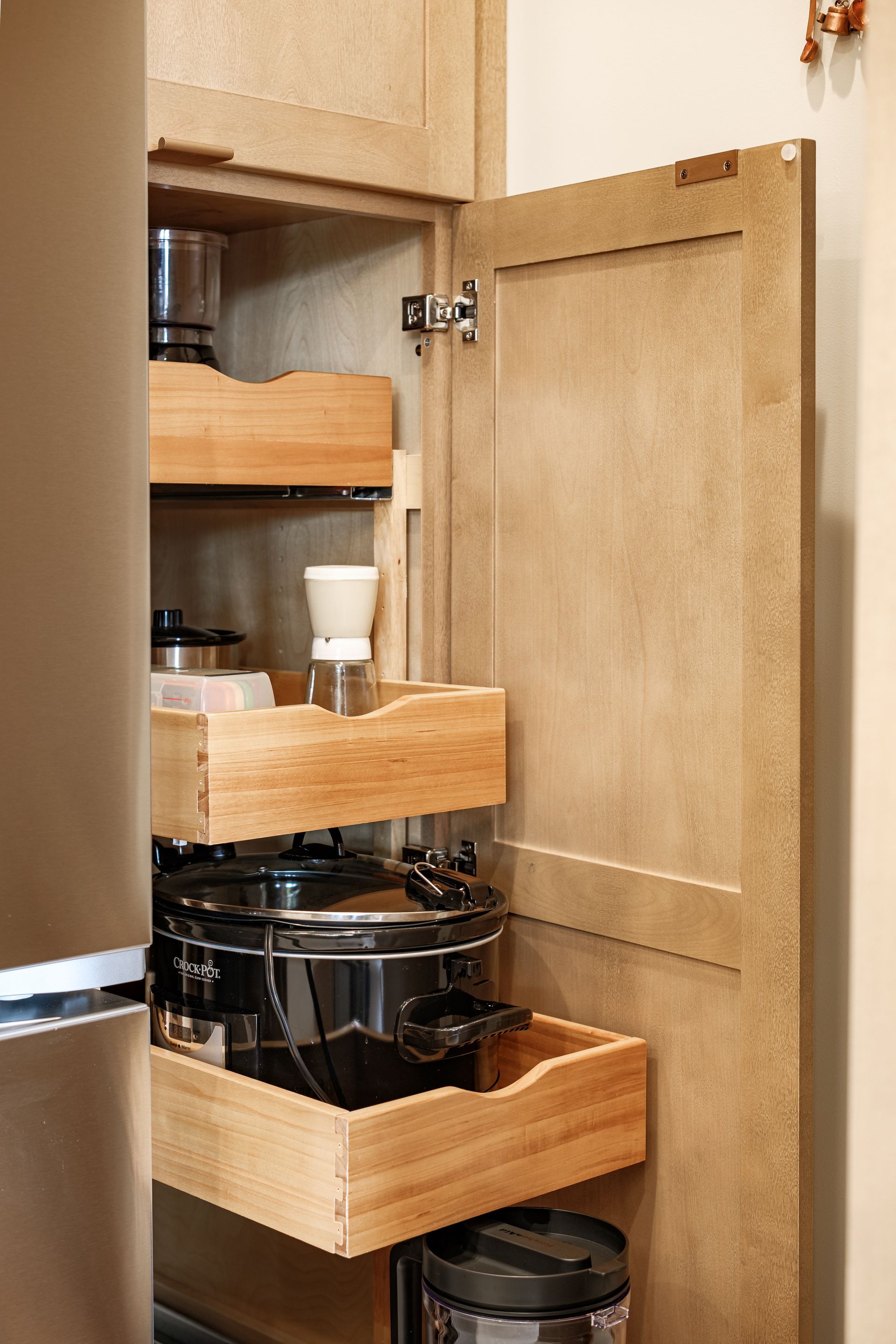Tall wooden kitchen cabinet with pull-out shelves holding small appliances, next to a refrigerator.