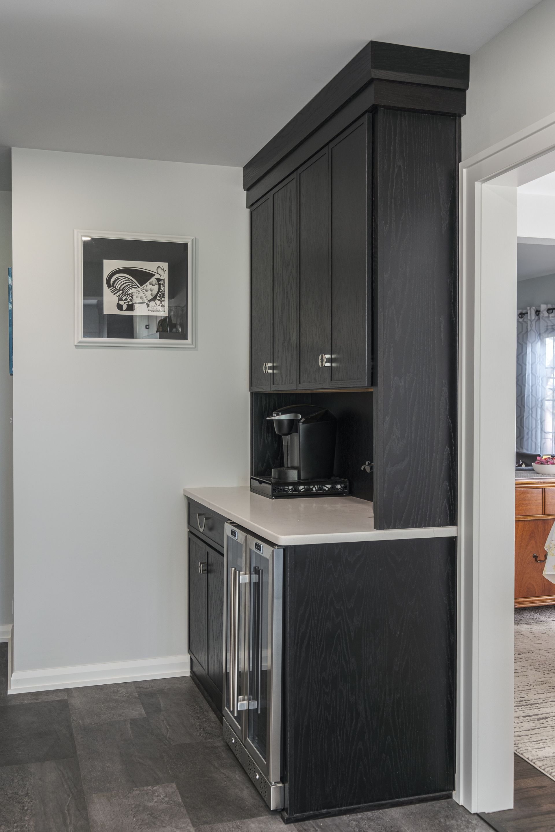 Dark cabinet coffee station with a mini-fridge, built into a narrow hallway next to a framed artwork.