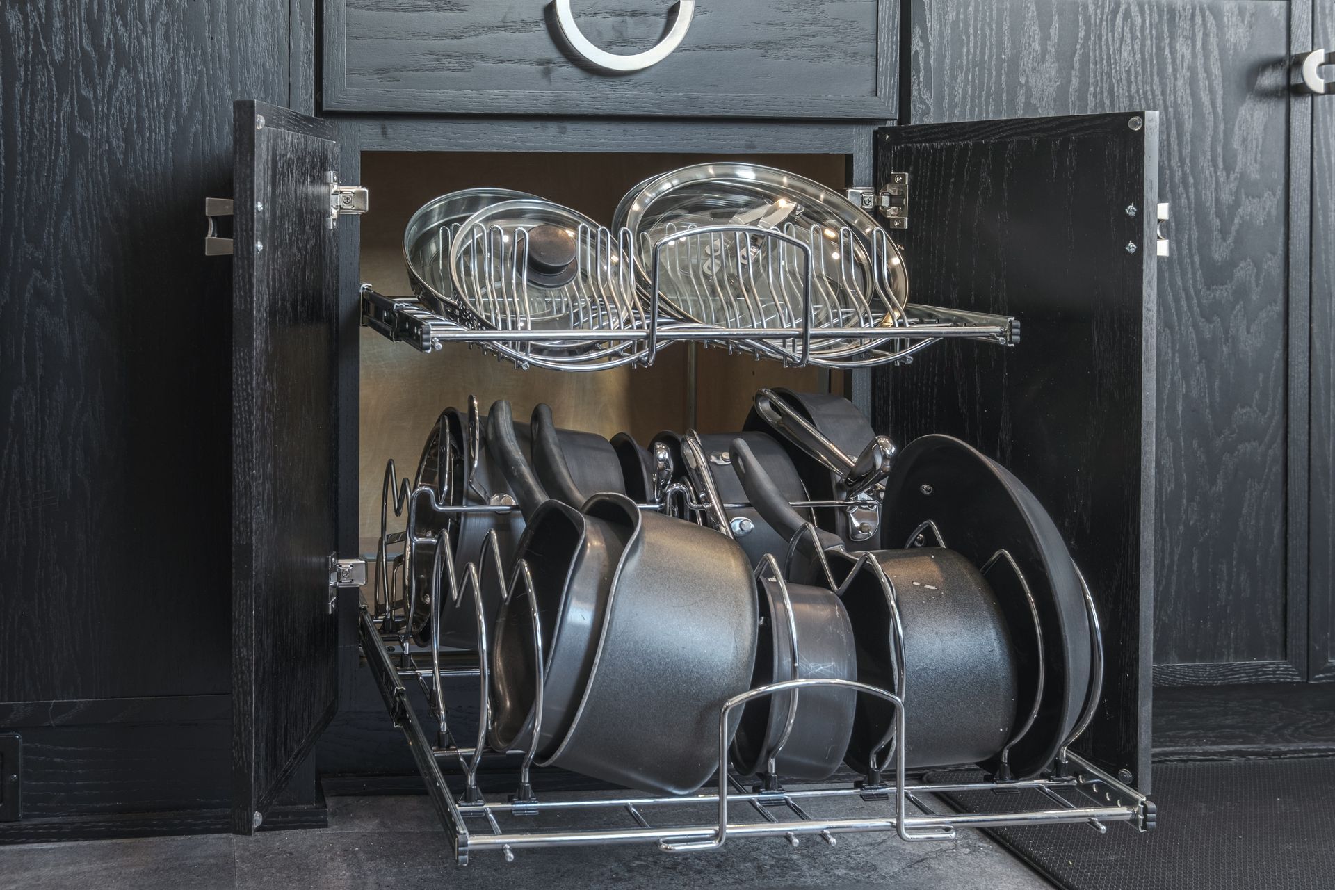 Black kitchen cabinet open to reveal a pull-out rack holding pots and pans, silver.