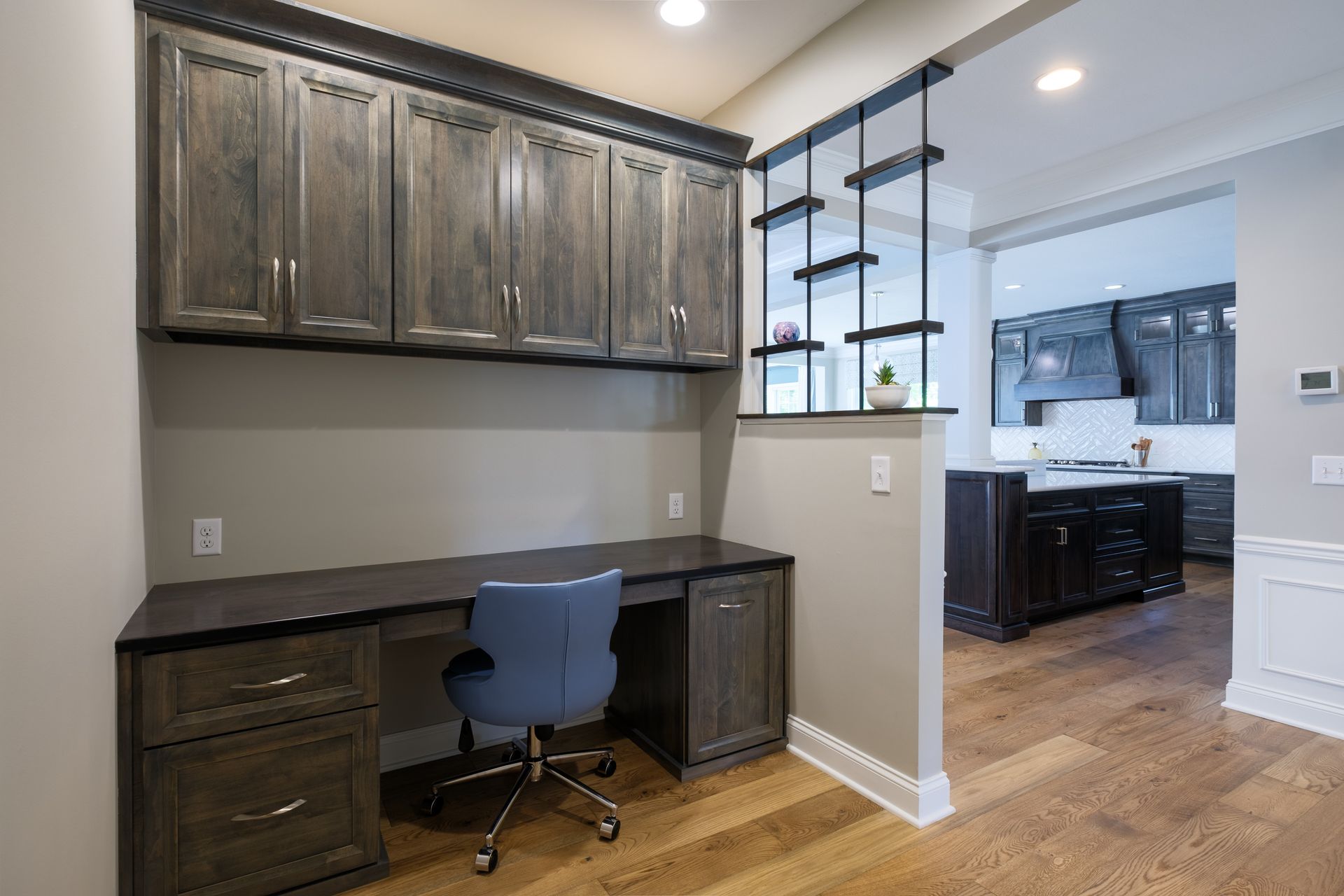 Home office with grey cabinetry, desk, and blue chair; kitchen in the background.
