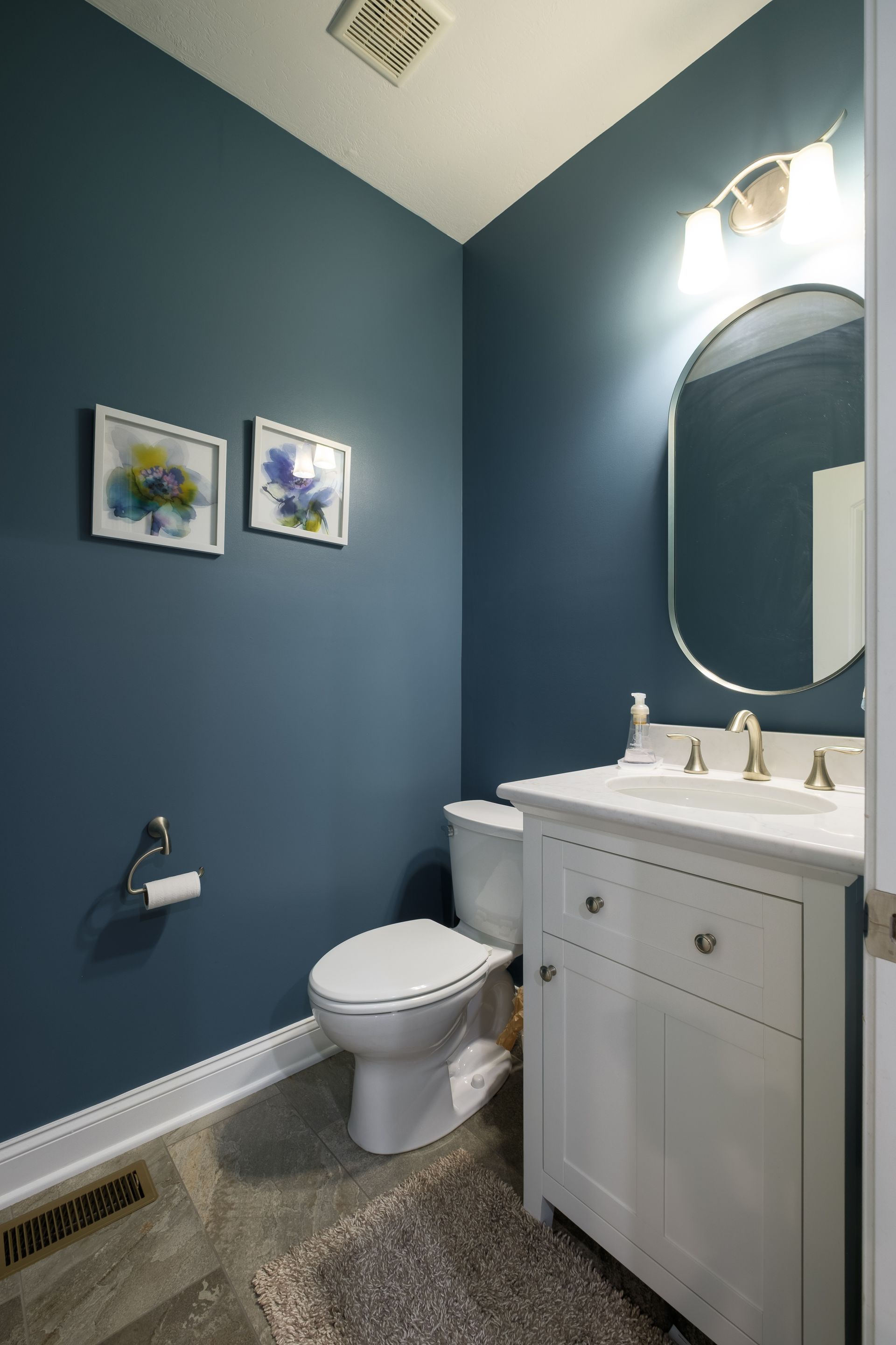 A small, blue-walled bathroom with white vanity, toilet, and artwork. A silver-framed mirror reflects the overhead light.