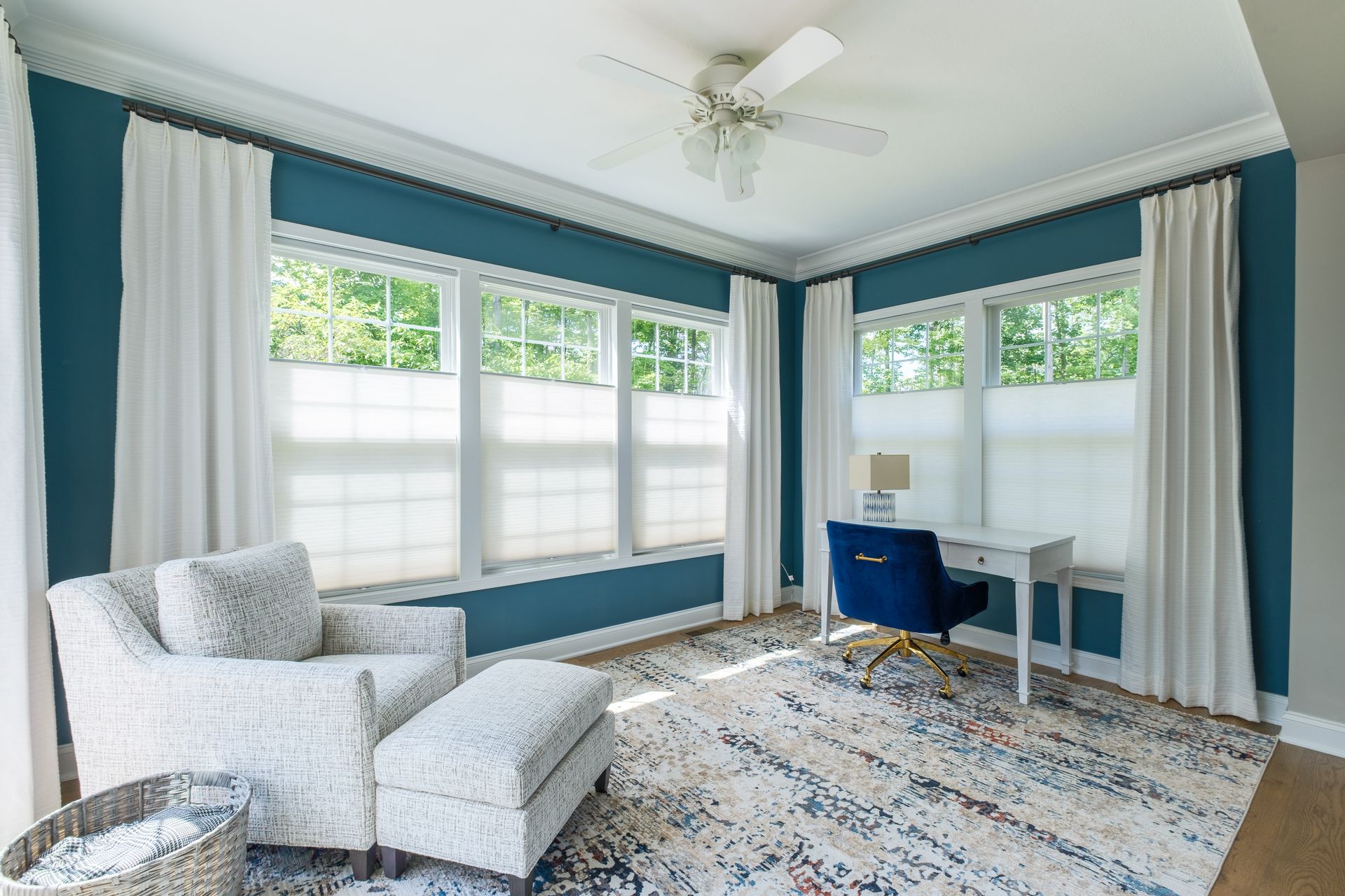 A bright room with blue walls, white curtains, a desk, chair, and armchair, with a patterned rug.