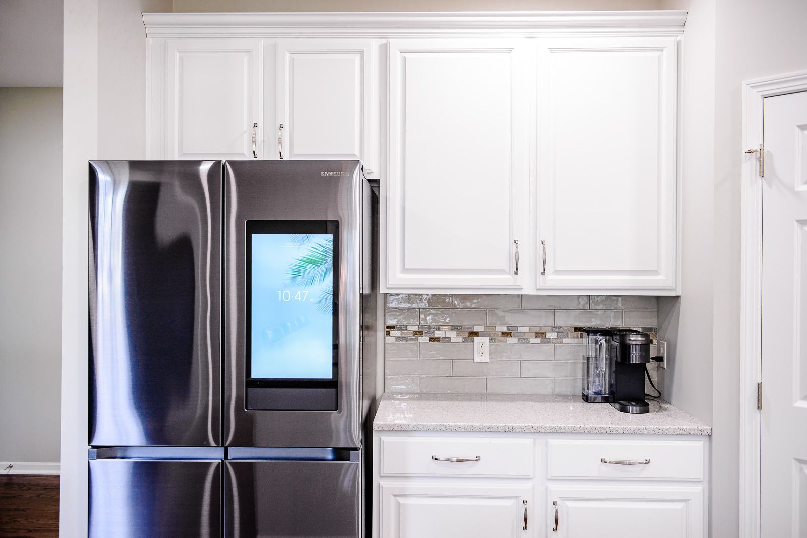 Stainless steel refrigerator with built-in screen, white cabinets, and countertop coffee maker in a kitchen.