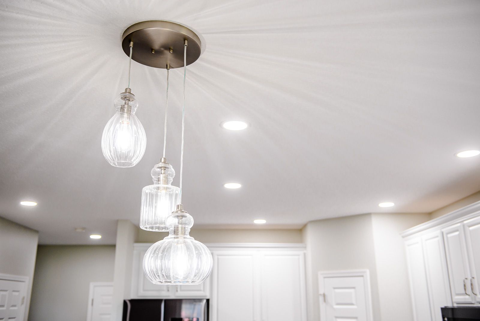Three glass pendant lights hang from a white ceiling. Recessed lighting illuminates a kitchen with white cabinets.